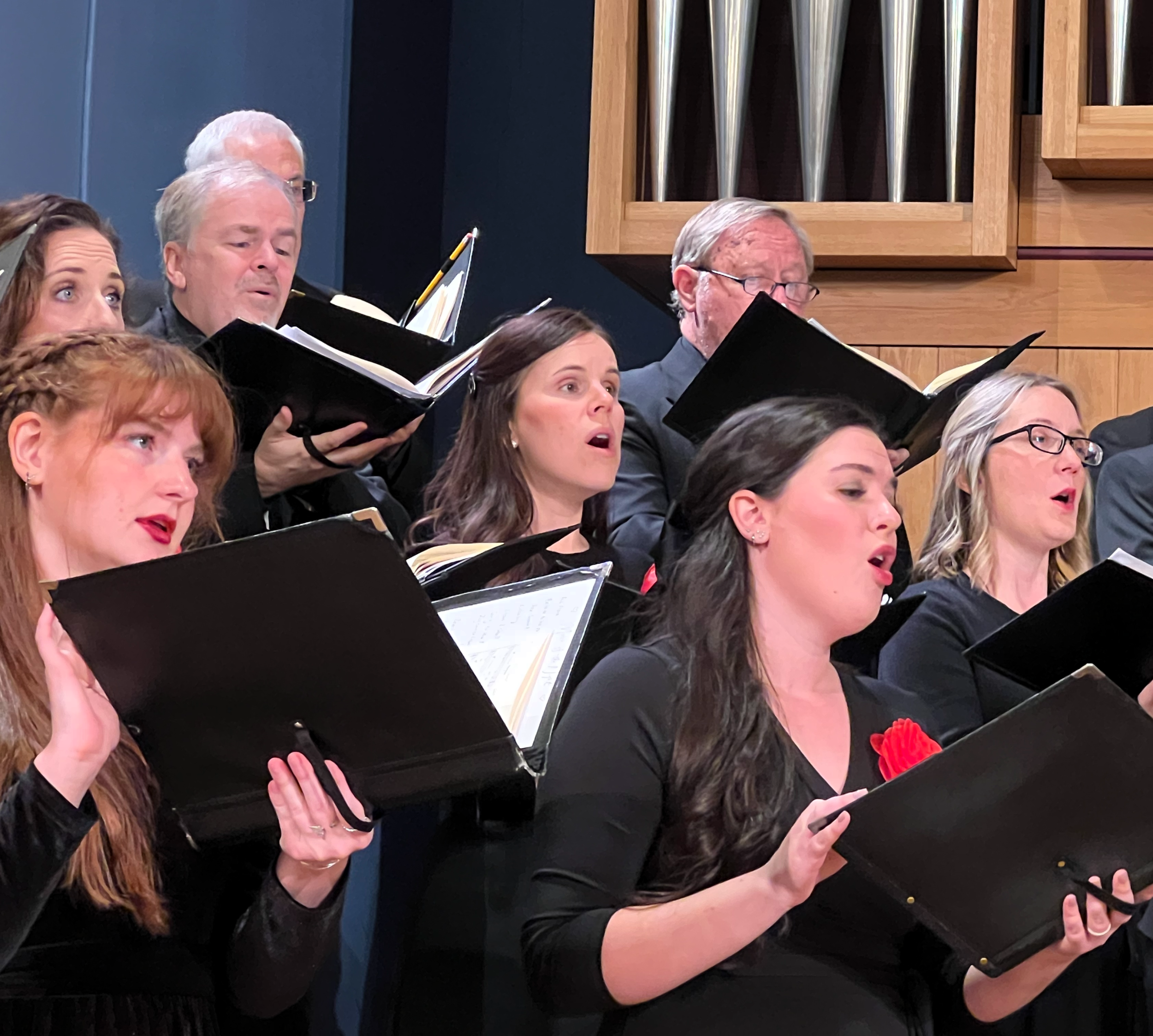A choir of men and women singing, holding sheet music, with some wearing black formal attire and red accents, in a concert hall with a wooden pipe organ in the background.