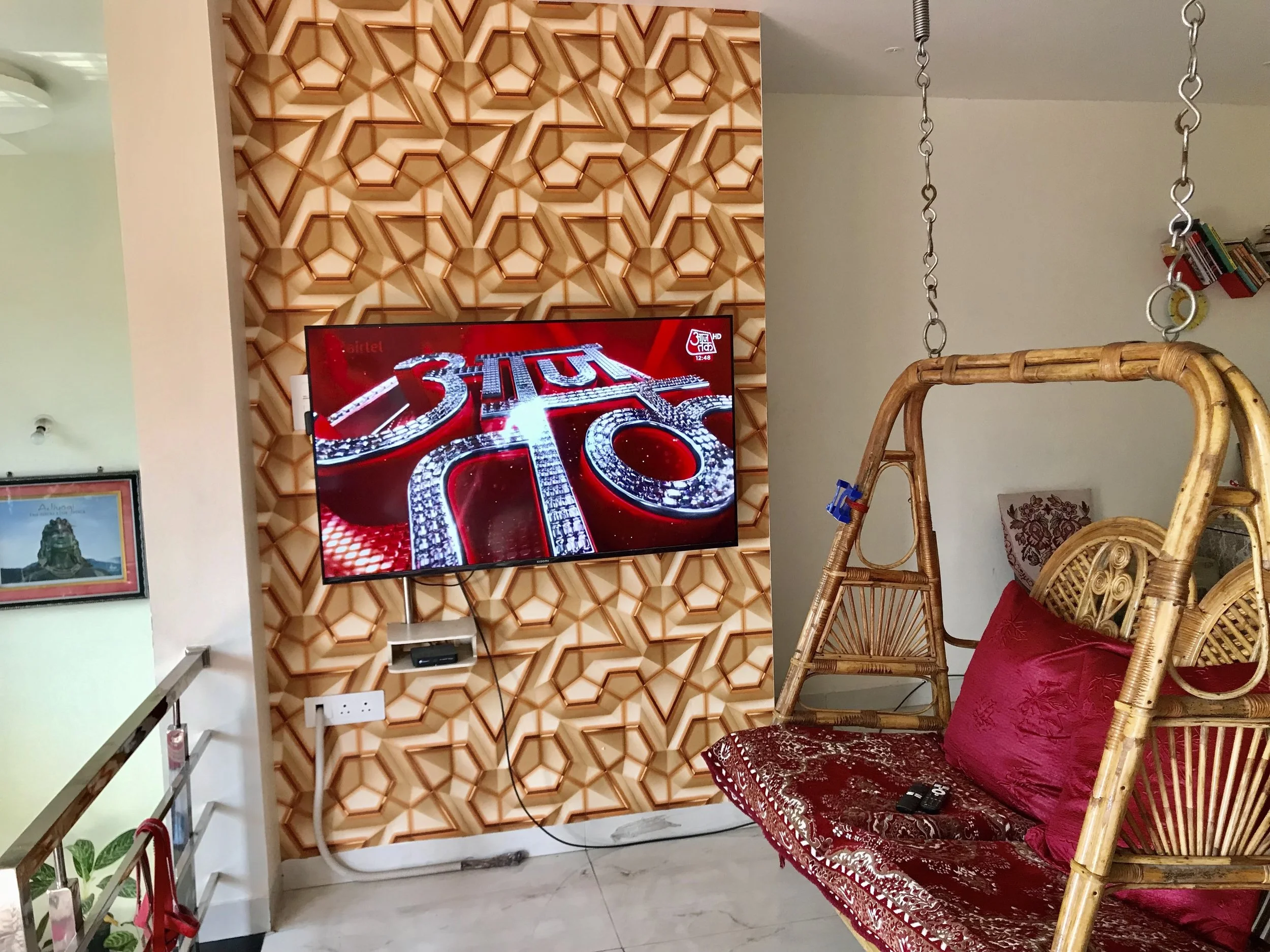 Interior of a living room with a flat screen TV on a wood-paneled wall with geometric patterns, wicker chairs, red cushions, and hanging chains. Part of a staircase with potted plants and artwork are also visible.