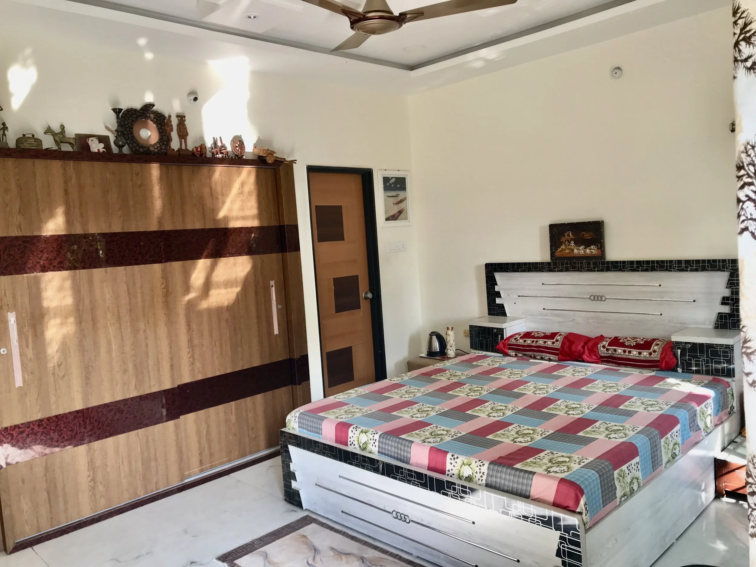 A bedroom with a colorful patchwork quilt on a bed, a wooden ceiling fan, a wooden cabinet with decorative items and small toys on top, a framed picture on the wall, a door, and a partially visible window with a black and white patterned curtain.