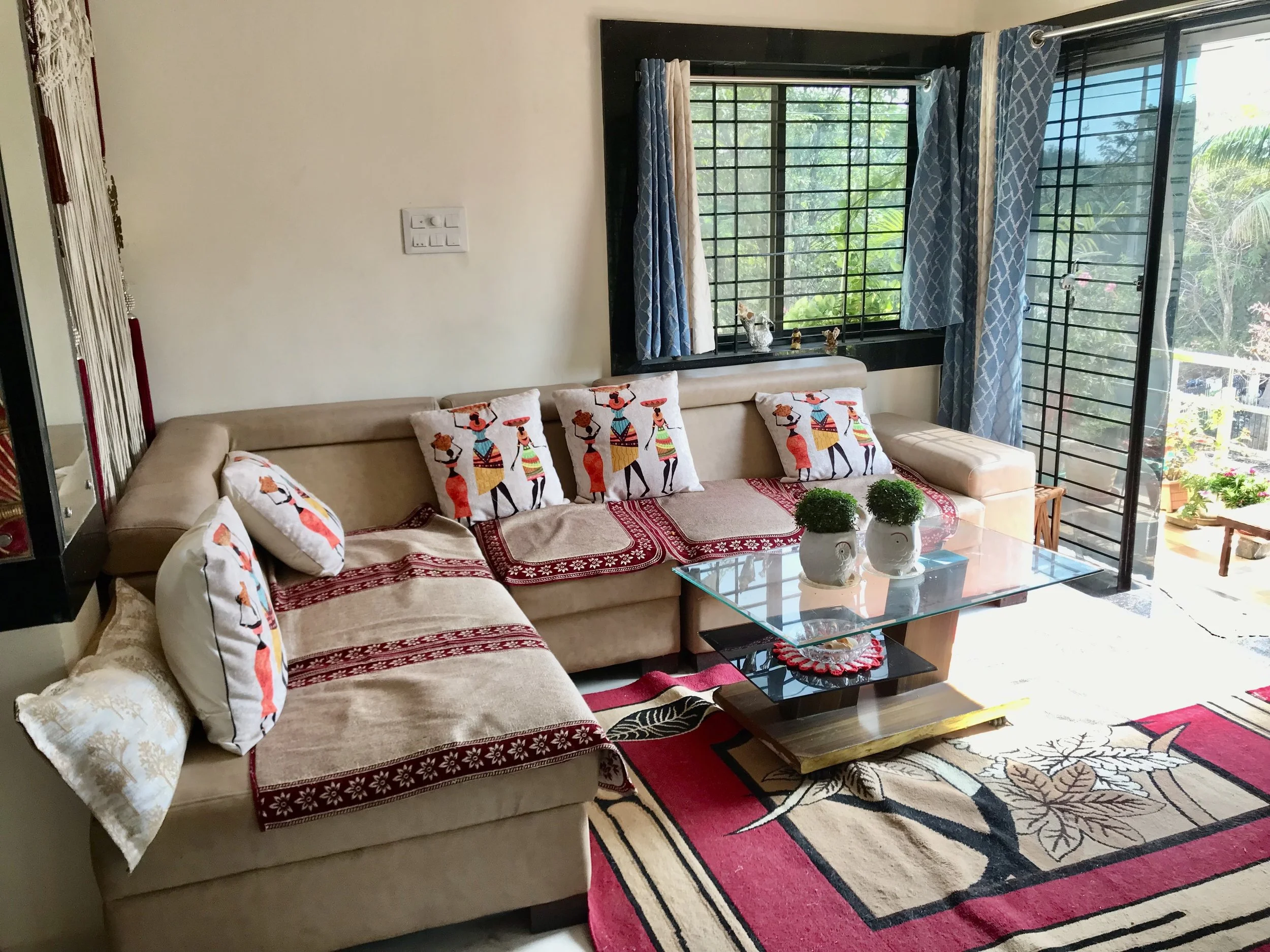 Living room with beige sofas decorated with colorful ethnic-patterned pillows, a glass coffee table, a patterned area rug, large windows with blue and white curtains, and indoor plants outside.