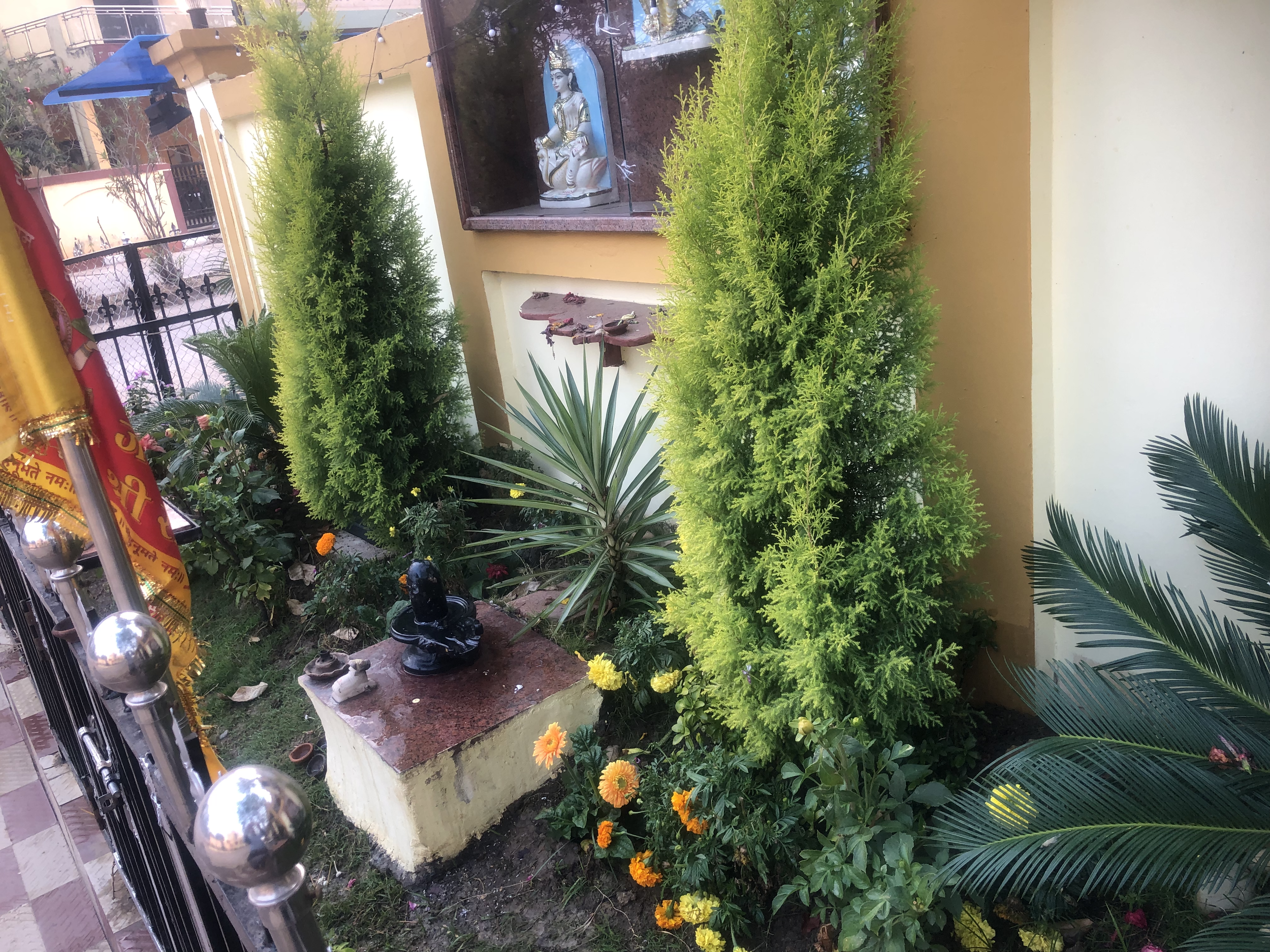 A small garden area with green bushes, flowering plants, and a small shrine or religious display featuring a statue of a deity, surrounded by decorations and objects, with a beige and yellow wall in the background.