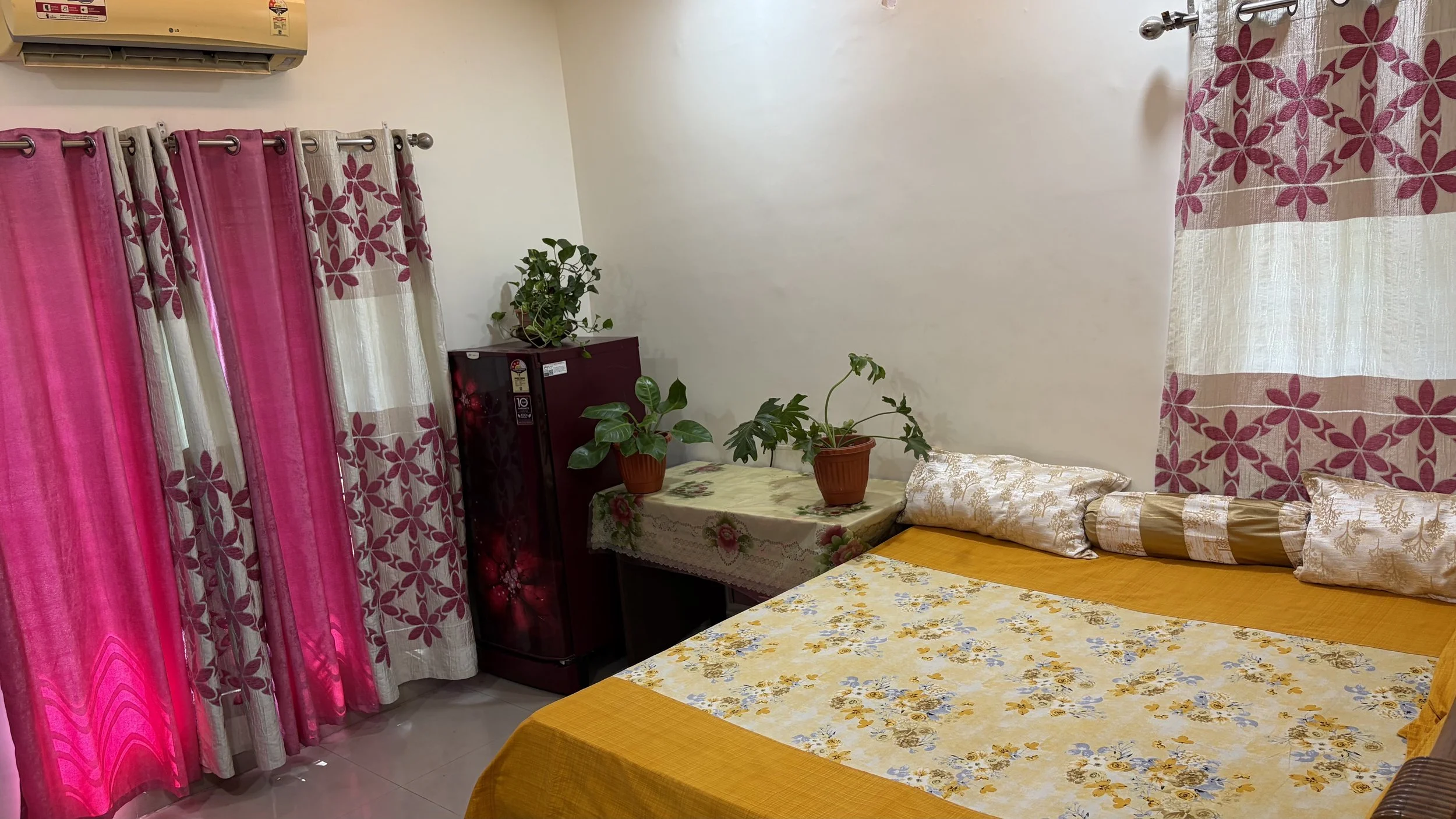 A cozy bedroom with pink and white floral curtains, a bed with a yellow and floral bedspread, decorative pillows, a table with potted plants, and an air conditioning unit on the wall.