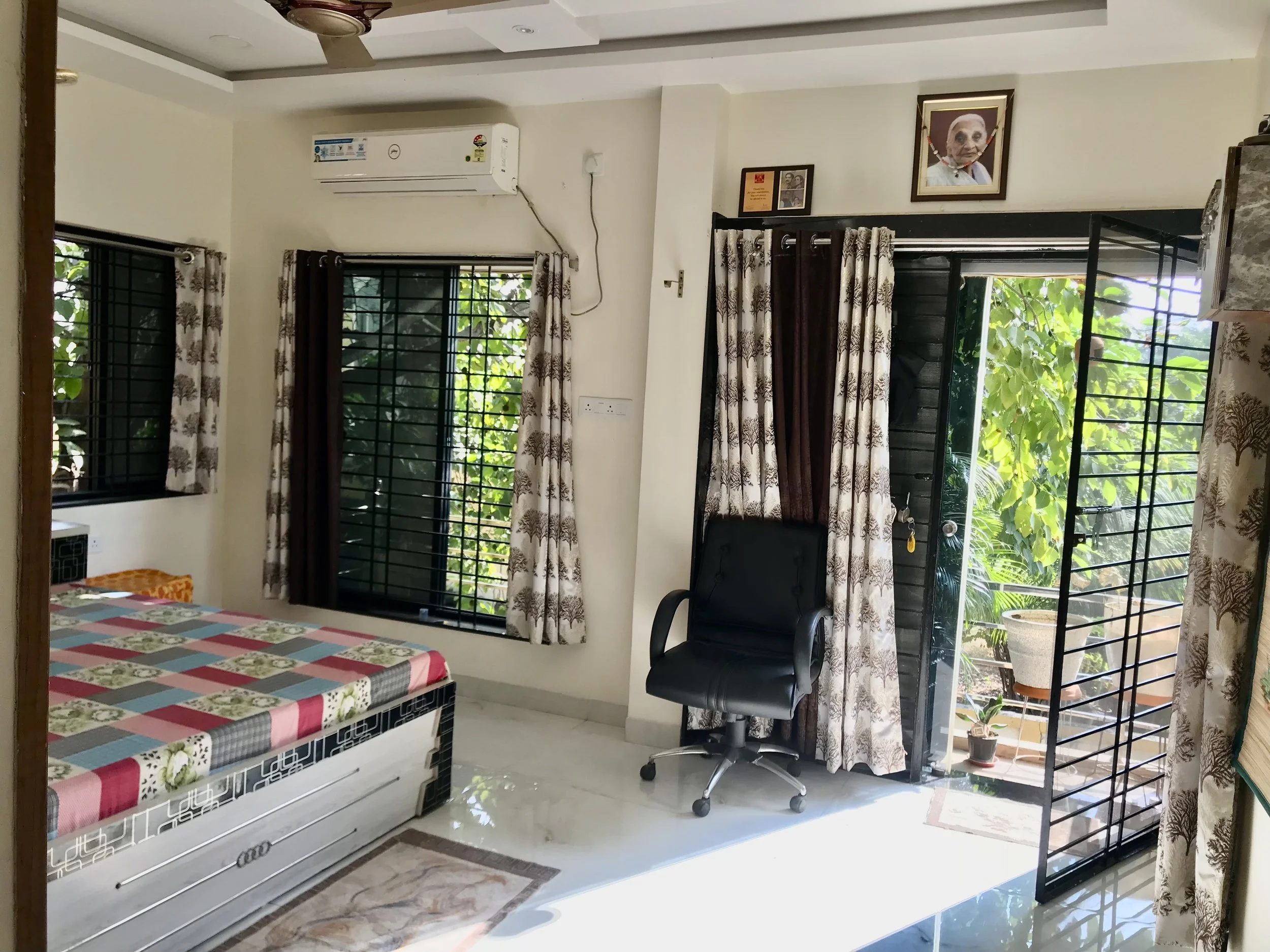 A room with two large windows with metal bars and floral curtains, a wall-mounted air conditioner, a framed portrait of an elderly woman, a framed certificate, a black office chair on wheels, and a table with a multicolored checkered tablecloth. There is also a door leading outside to a garden with green foliage.