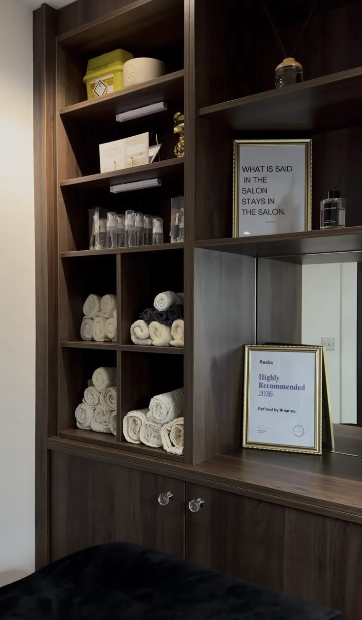 Wooden shelf unit with rolled towels, bottles, and framed signs, in a salon or spa setting.