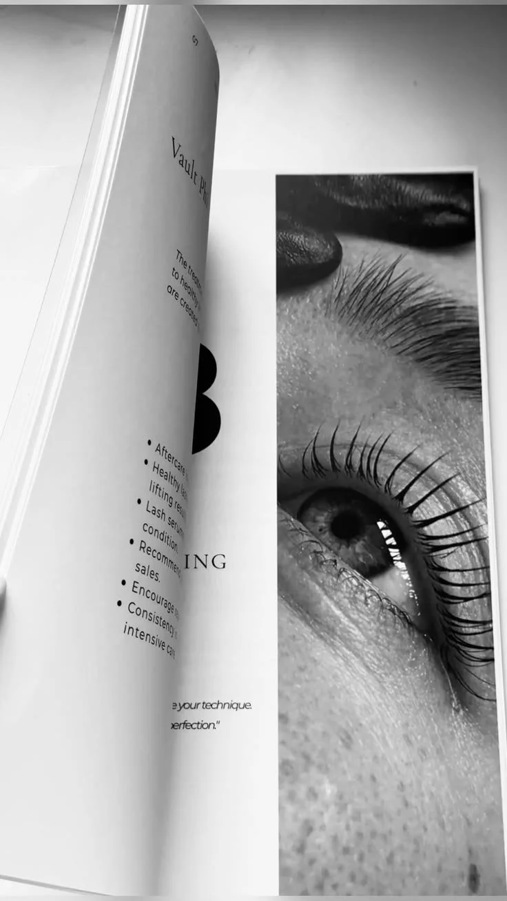 A close-up black and white photograph of a person's eye with long eyelashes, part of a magazine or book page visible next to it displaying text and a large black letter 'B'.