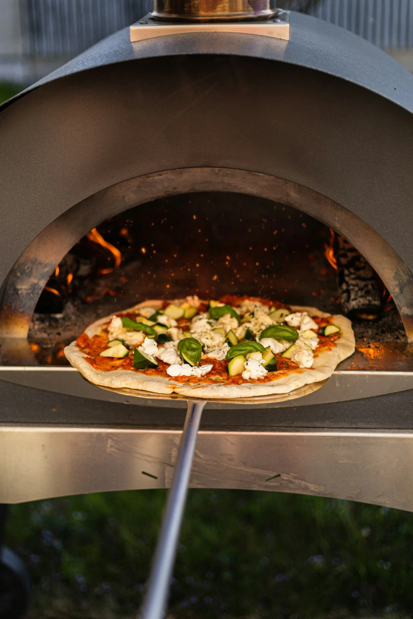 A pizza baking in an open hearth pizza oven, showing toppings like basil, mushrooms, and zucchini.