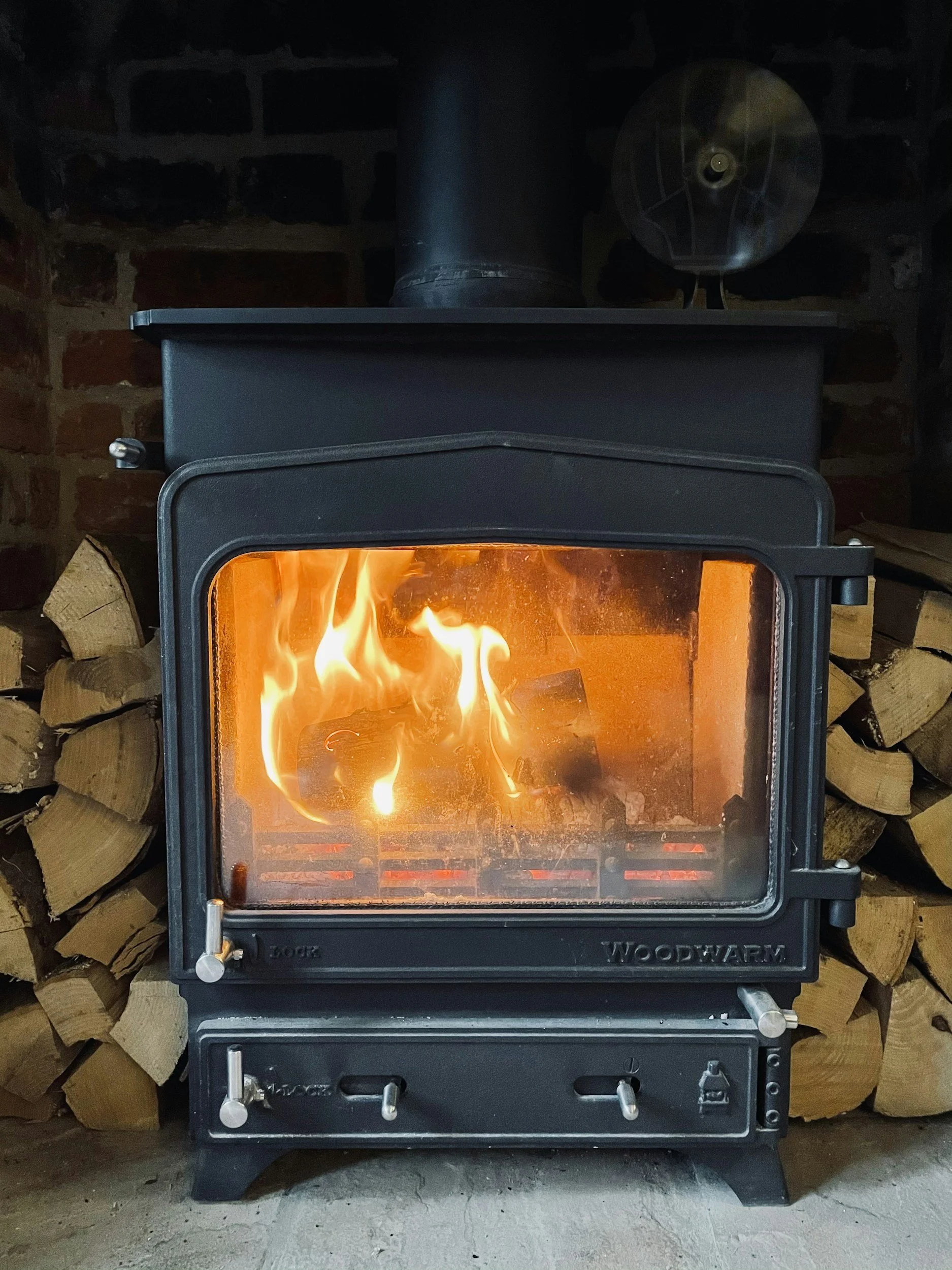 Woodstove with a fire inside, stacked firewood behind it.