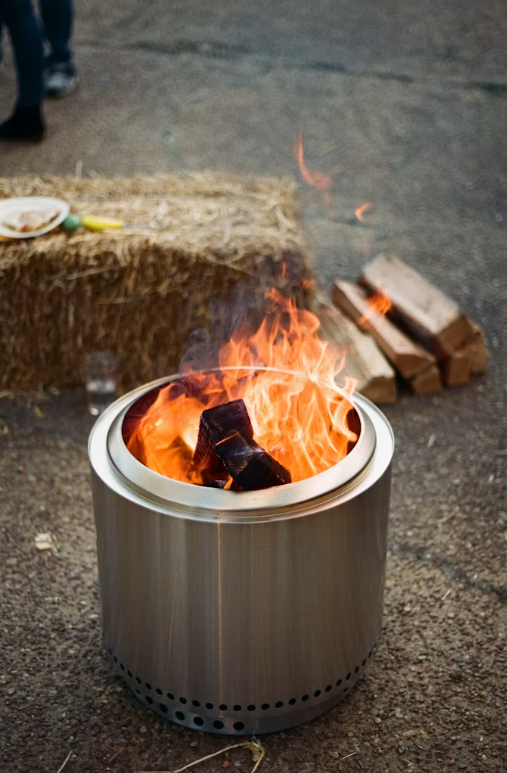 A modern metal fire pit with burning logs on a paved patio, with firewood in the background.