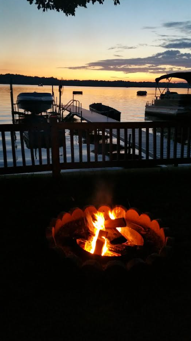 A lakeside scene at sunset with a fire pit in the foreground and boats docked at a pier in the background.