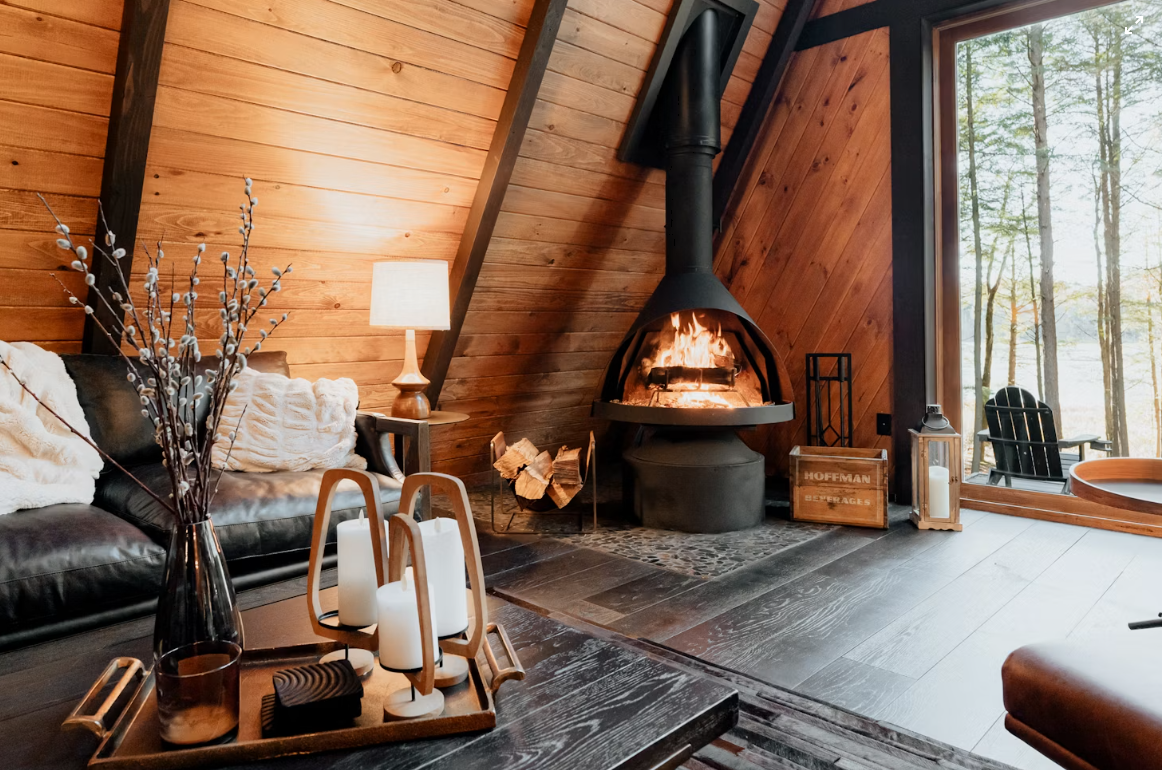Cozy living room with a wood-paneled wall, a lit fireplace, and a large window overlooking an outdoor view of trees and water. Includes leather sofa, table with candles, and decorative items.