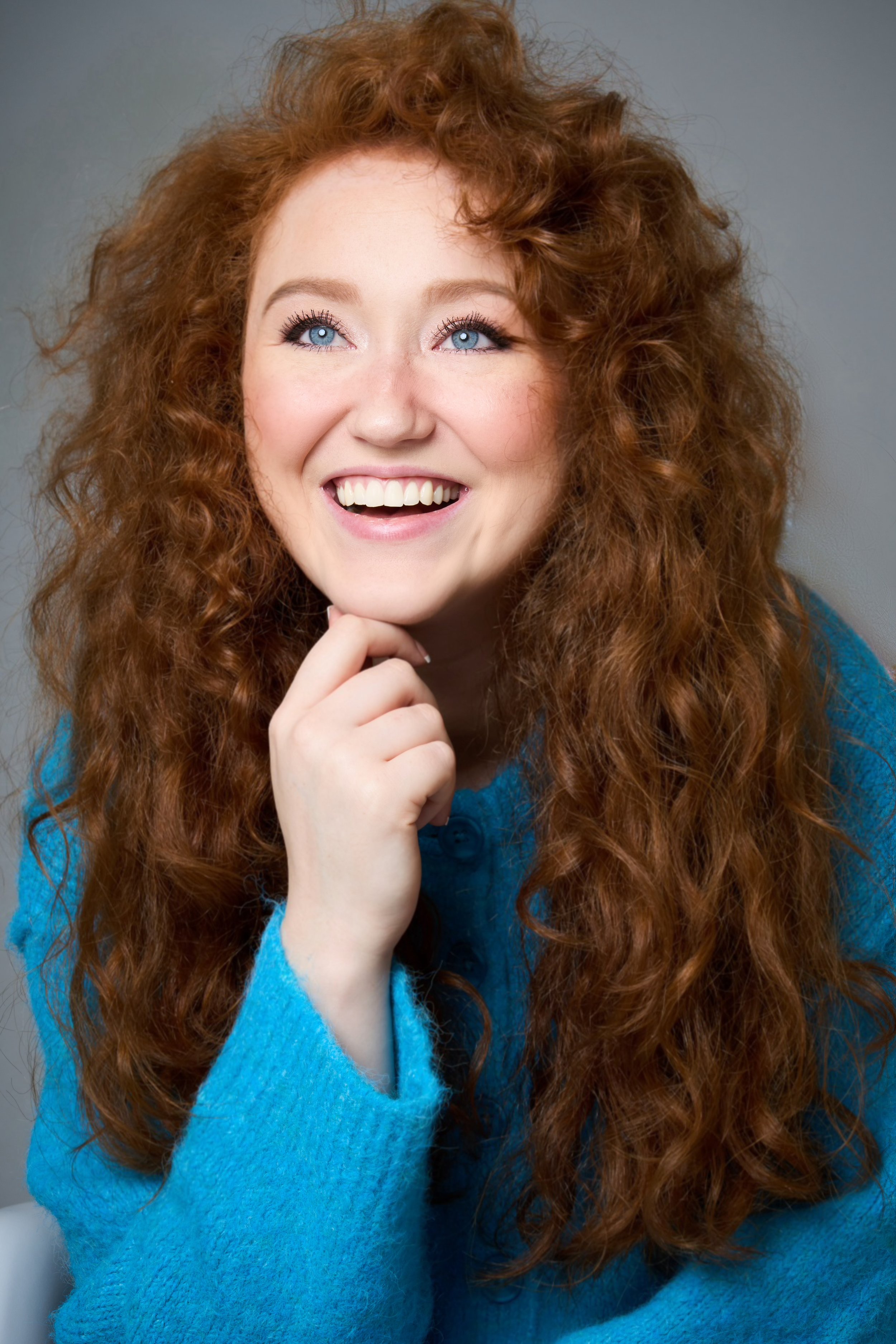 A woman with long, curly red hair and blue eyes smiling while resting her chin on her hand, wearing a blue sweater.