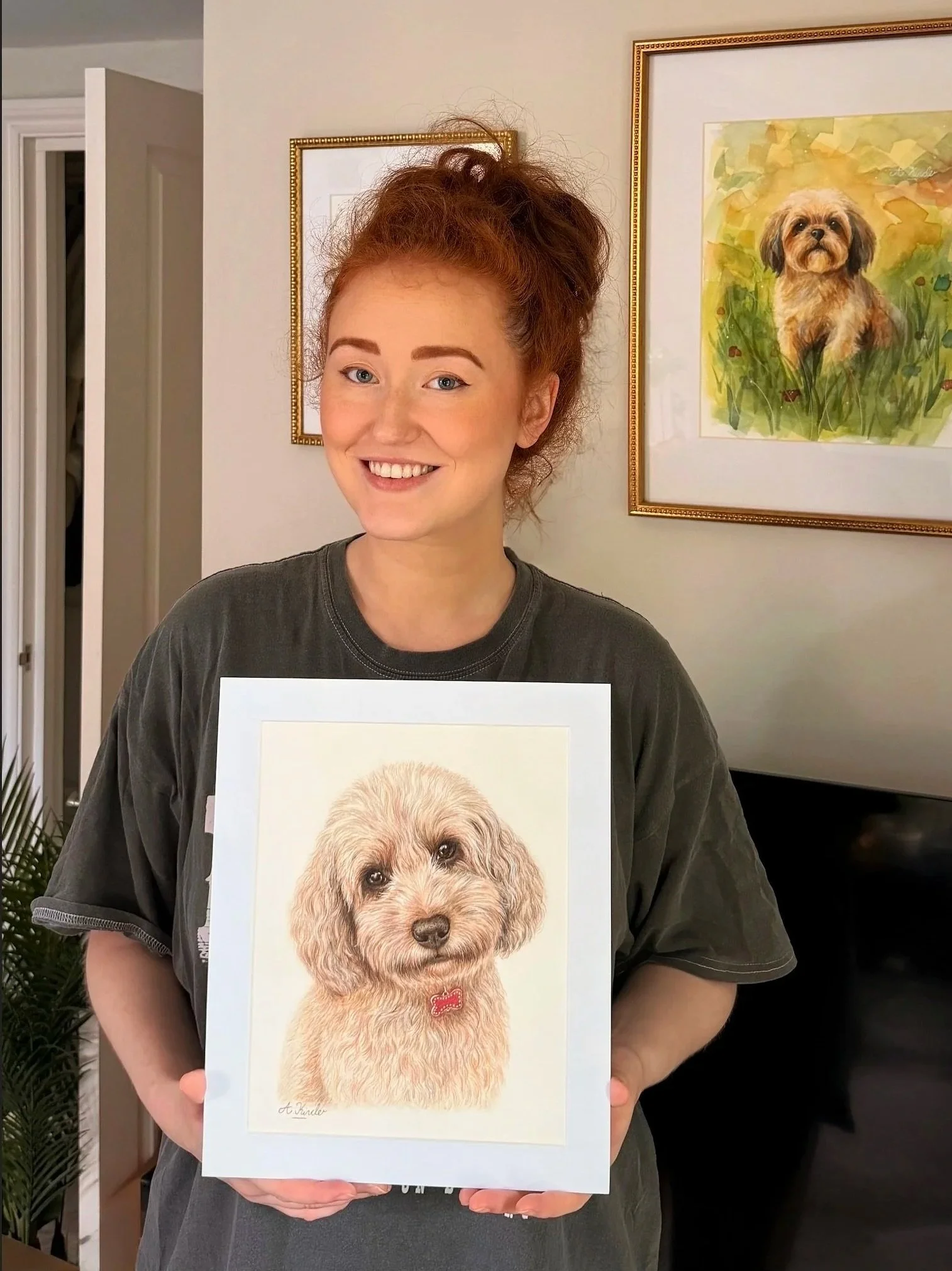 A woman with red curly hair smiling while holding a portrait of a beige dog with a red bow tie. Behind her, there is a framed watercolor painting of a similar dog on the wall.
