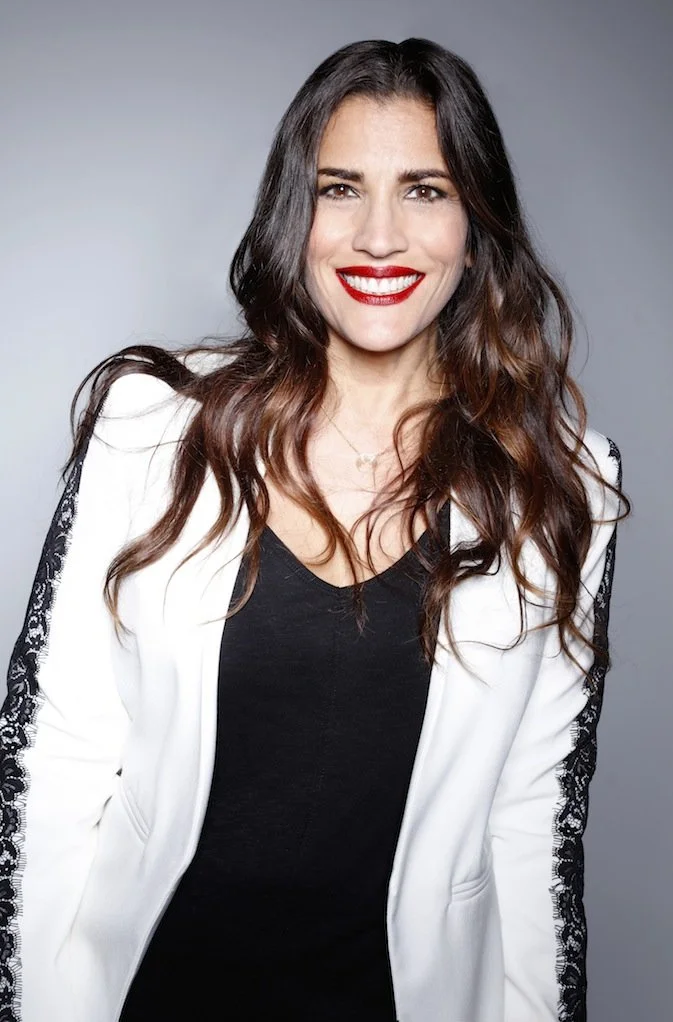 A woman with long brown hair, red lipstick, and a bright smile, wearing a white blazer with black lace trim over a black top, standing against a neutral gray background.