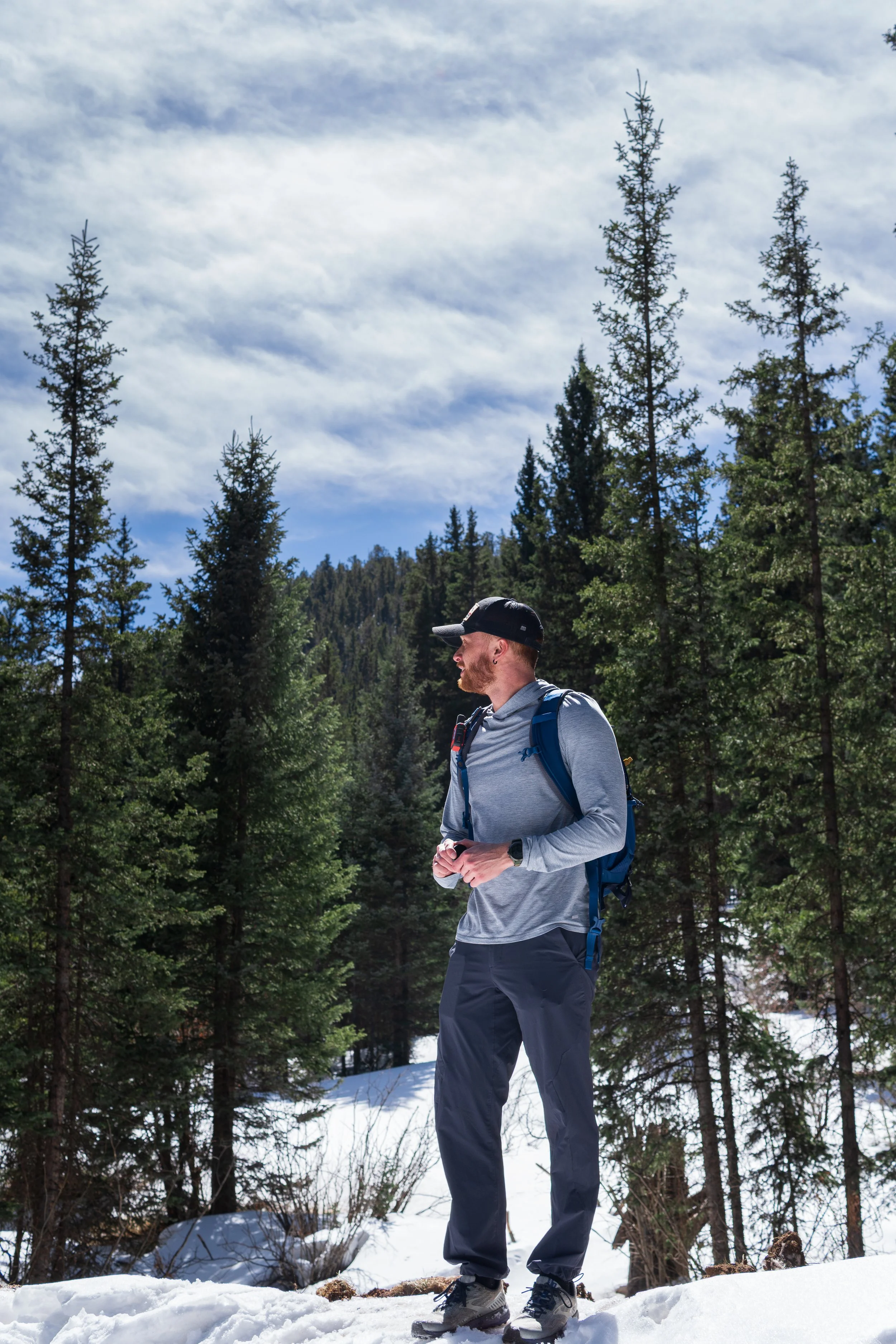Man hiking in a snowy forest with tall evergreen trees under a partly cloudy sky.