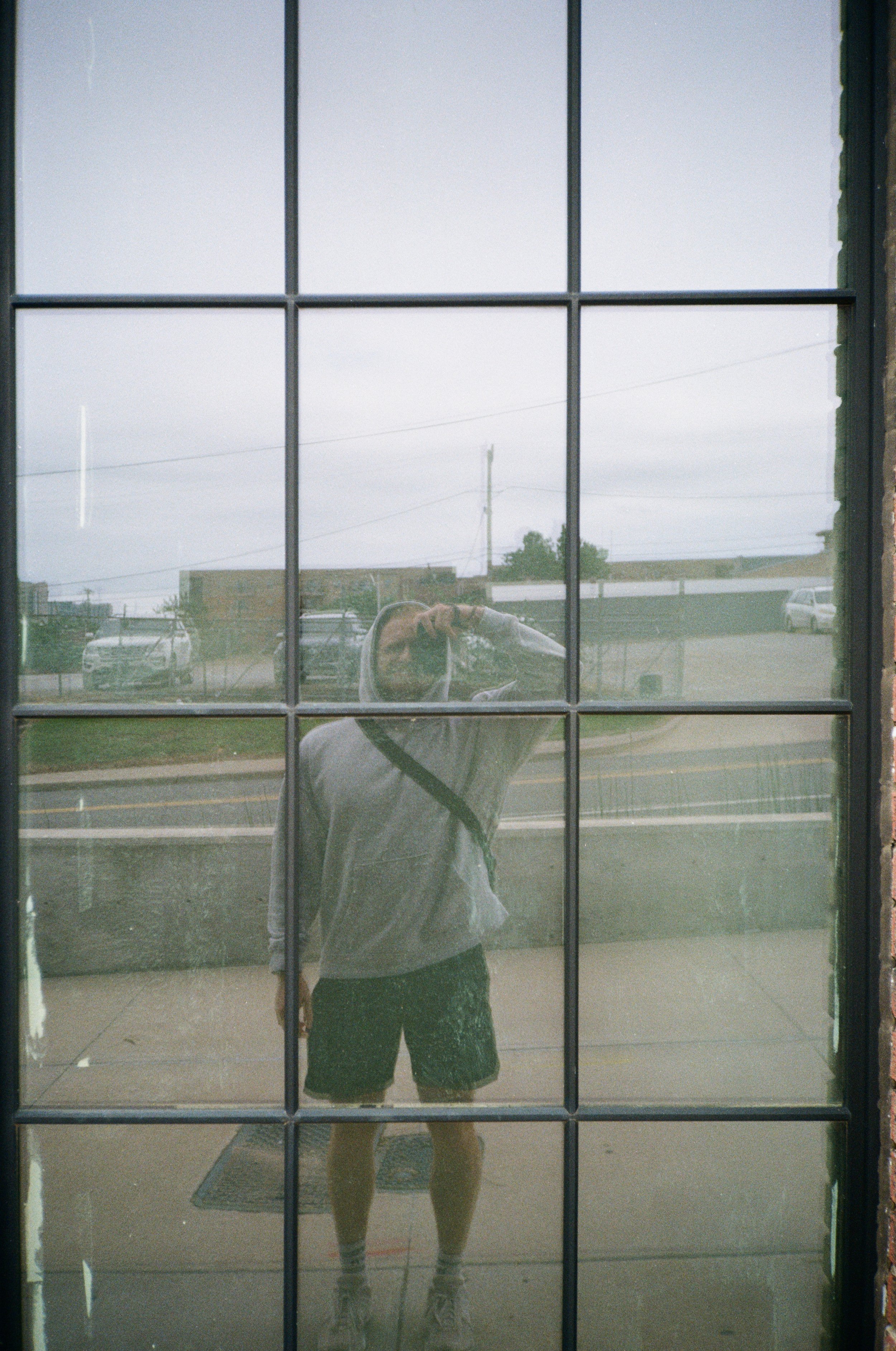 Reflection of a man taking a photo in a large window with multiple panes, showing an outdoor scene with cars, power lines, and cloudy sky.