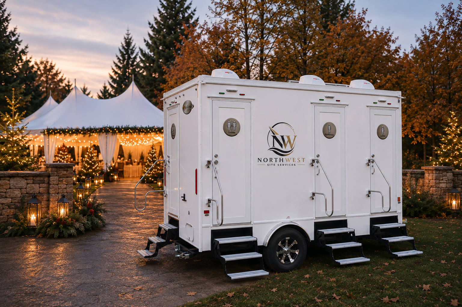 Portable restroom trailer parked outside a decorated outdoor event space with string lights, trees, and a white tent at sunset.