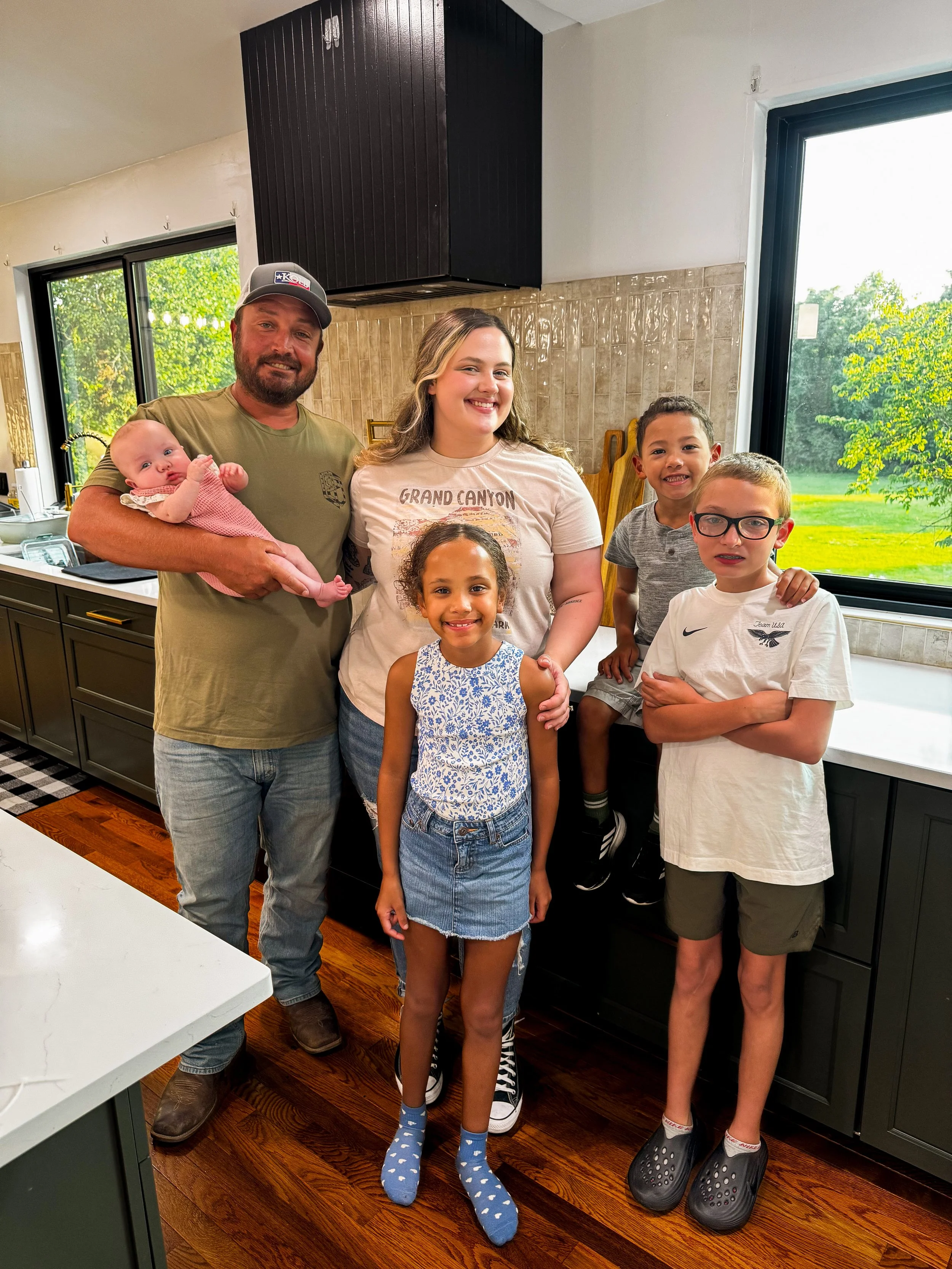A family of six standing in a kitchen with green cabinets and a large window. The man on the left is holding a baby. The woman in the middle and four children are smiling for the photo.