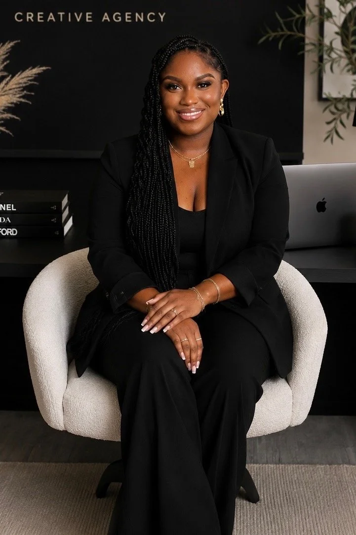 Owner of Creative agency in San Antonio Texas . A woman with long braided hair wearing a black blazer and pants, gold jewelry, sitting on a cream-colored chair in an office setting.