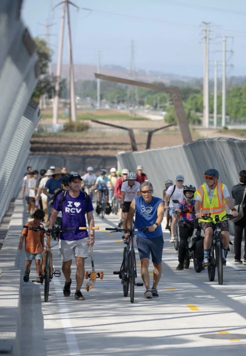 Group of people walking and cycling on the new Jeffrey Open Space Train bridge with metal railings and utility poles in the background. Jeremy Ficarola walks in front wearing his I-heart-RCV tee. Vote YES on Measure D for RCV!