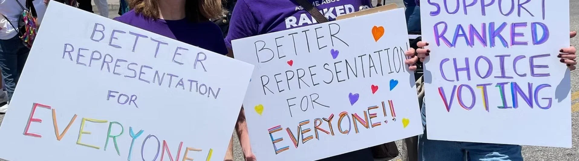 People holding signs with messages supporting ranked choice voting, featuring colorful handwritten text and decorative hearts. Vote YES on Measure D for RCV!