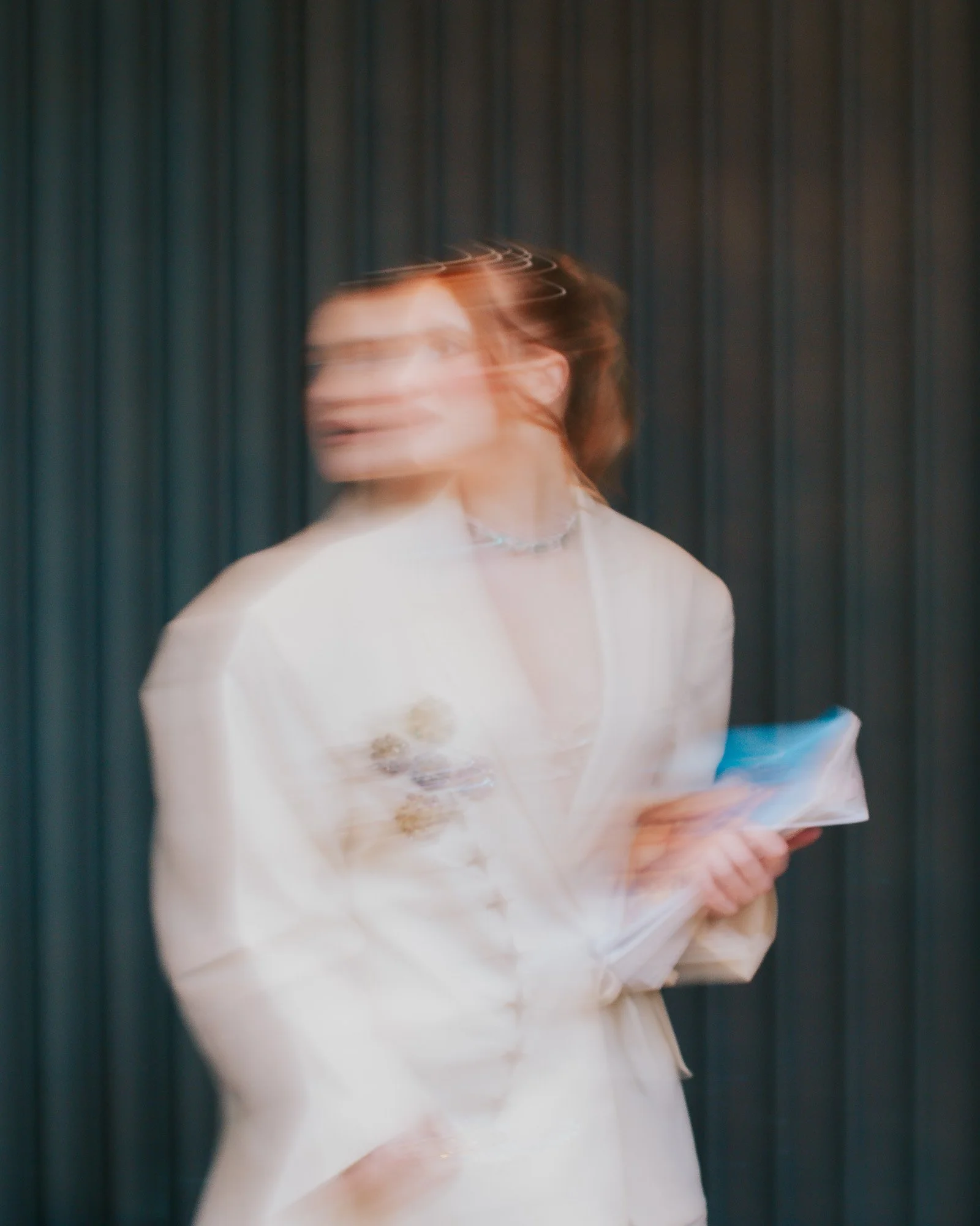 A woman with brown hair wearing glasses, a beige top, and a pearl necklace, holding a tray and standing in front of a dark curtain.