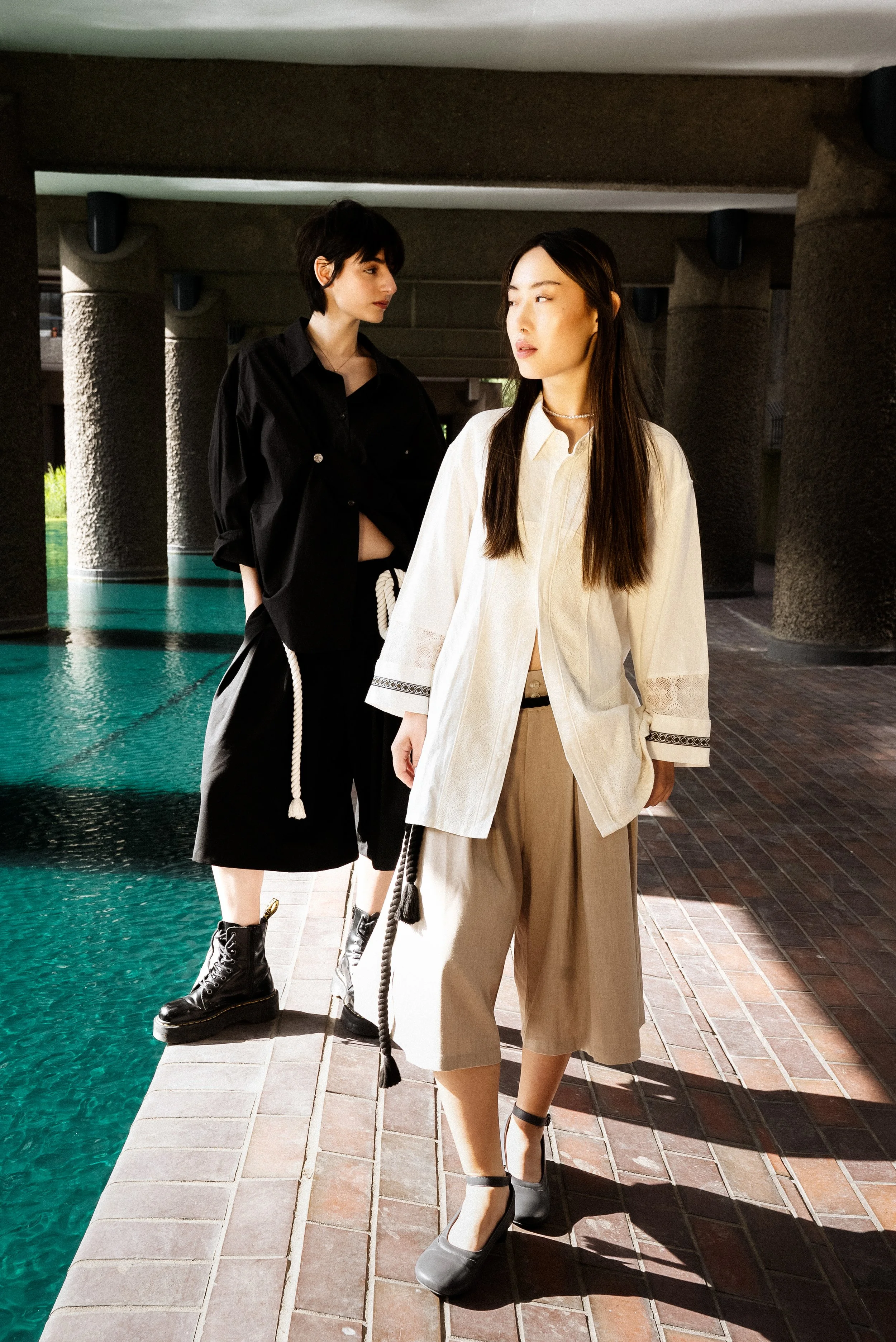 Two women standing by a pool under a concrete structure, dressed in trendy high fashion clothing.