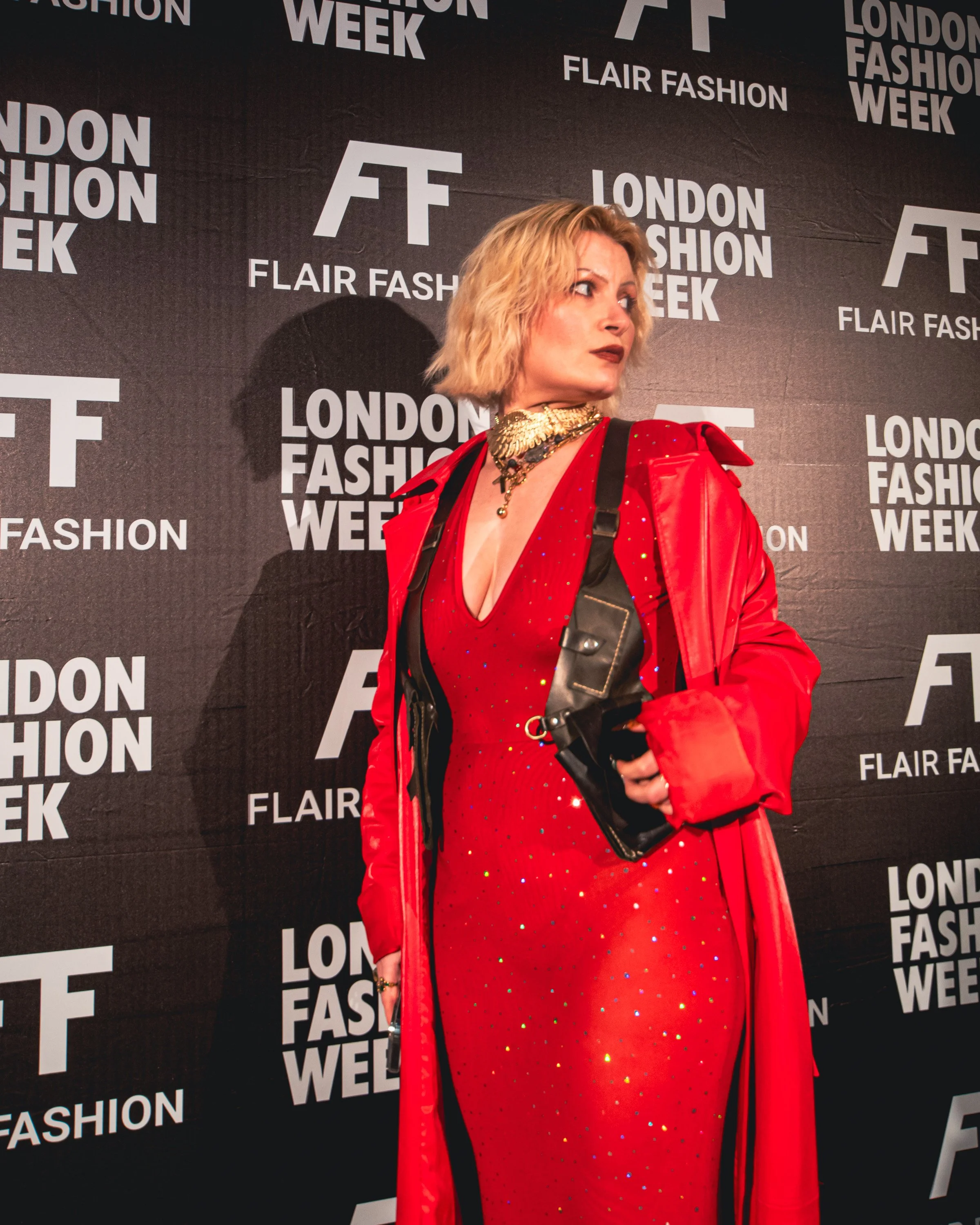 A woman with blonde hair wearing a red dress, red jacket, and gold jewelry standing against a background with 'London Fashion Week' and 'Flair Fashion' logos.