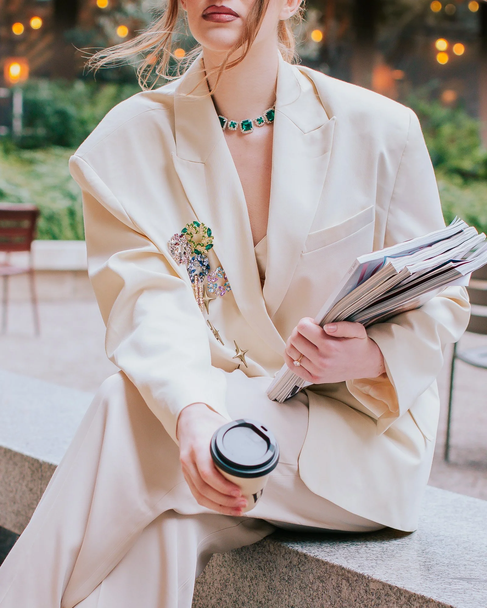 A woman holding a disposable coffee cup, wearing a beige blazer, a green gemstone necklace, and a brooch, sitting outdoors with blurred background lights.