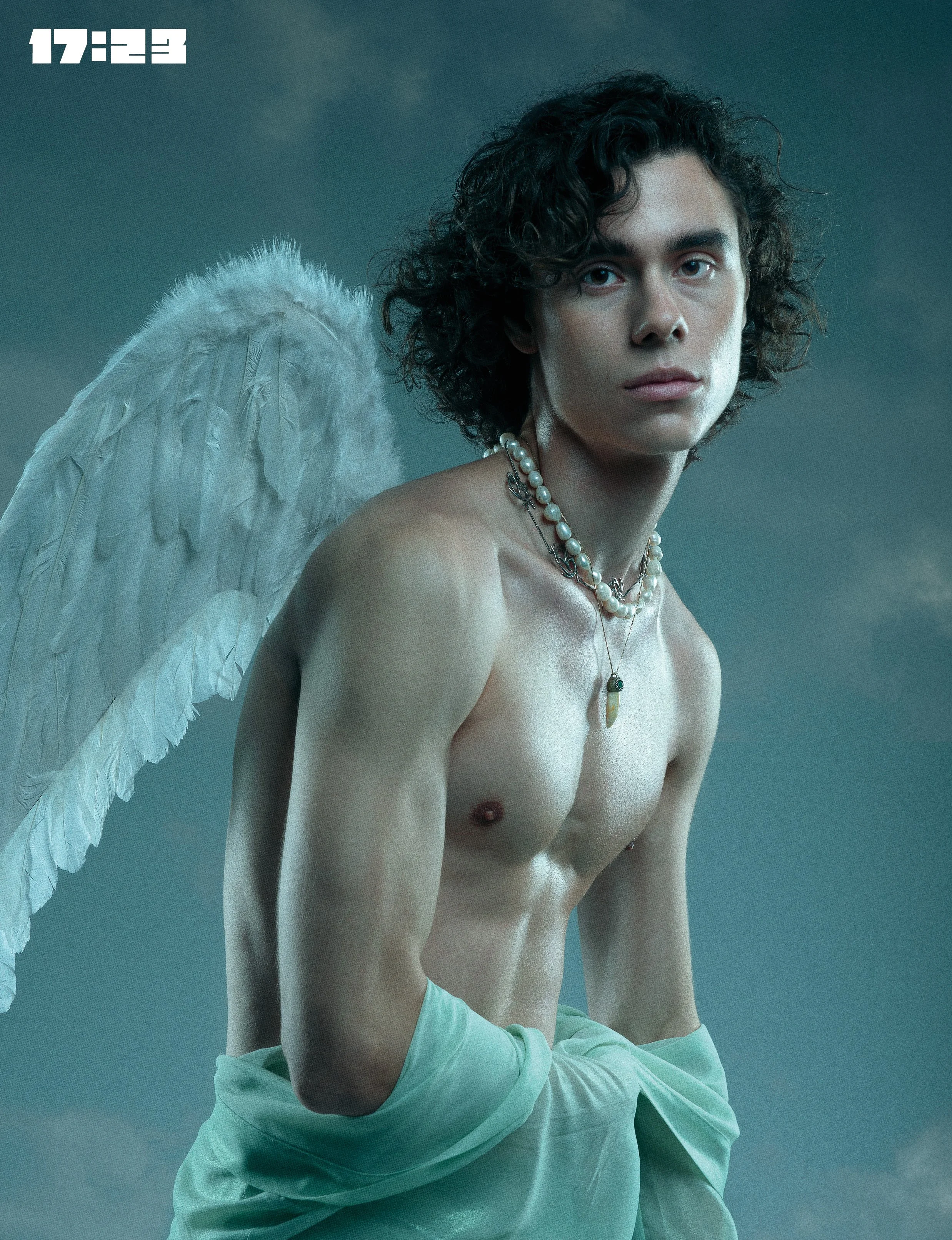 A young man with curly dark hair and piercing eyes, shirtless, wearing layered necklaces, including pearls, against a muted teal background. A large white feathered angel wing is visible behind him on the left side of the image. The time 17:23 is displayed in the top left corner.