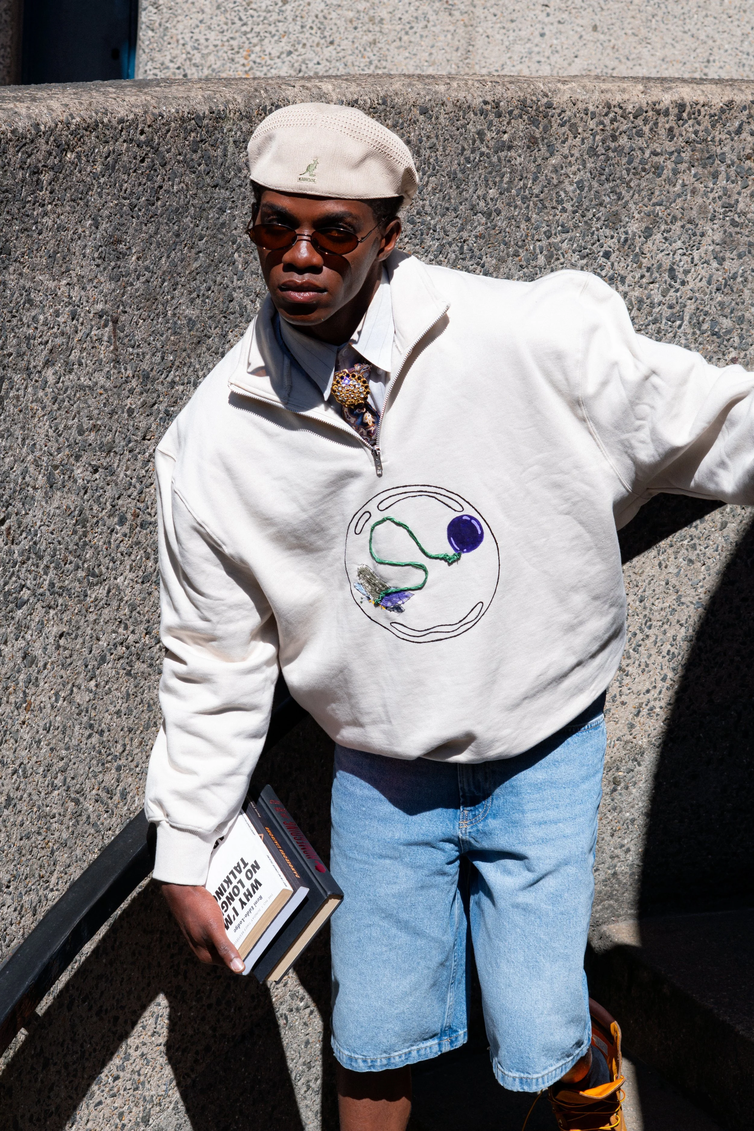 A person wearing a white sweatshirt with a circular NASA logo, blue denim shorts, orange hiking shoes, a beige cap with a dinosaur logo, and sunglasses, stands against a concrete wall. They are holding a book titled 'WHY NO LOGIC TALKS' and a black case in their left hand.