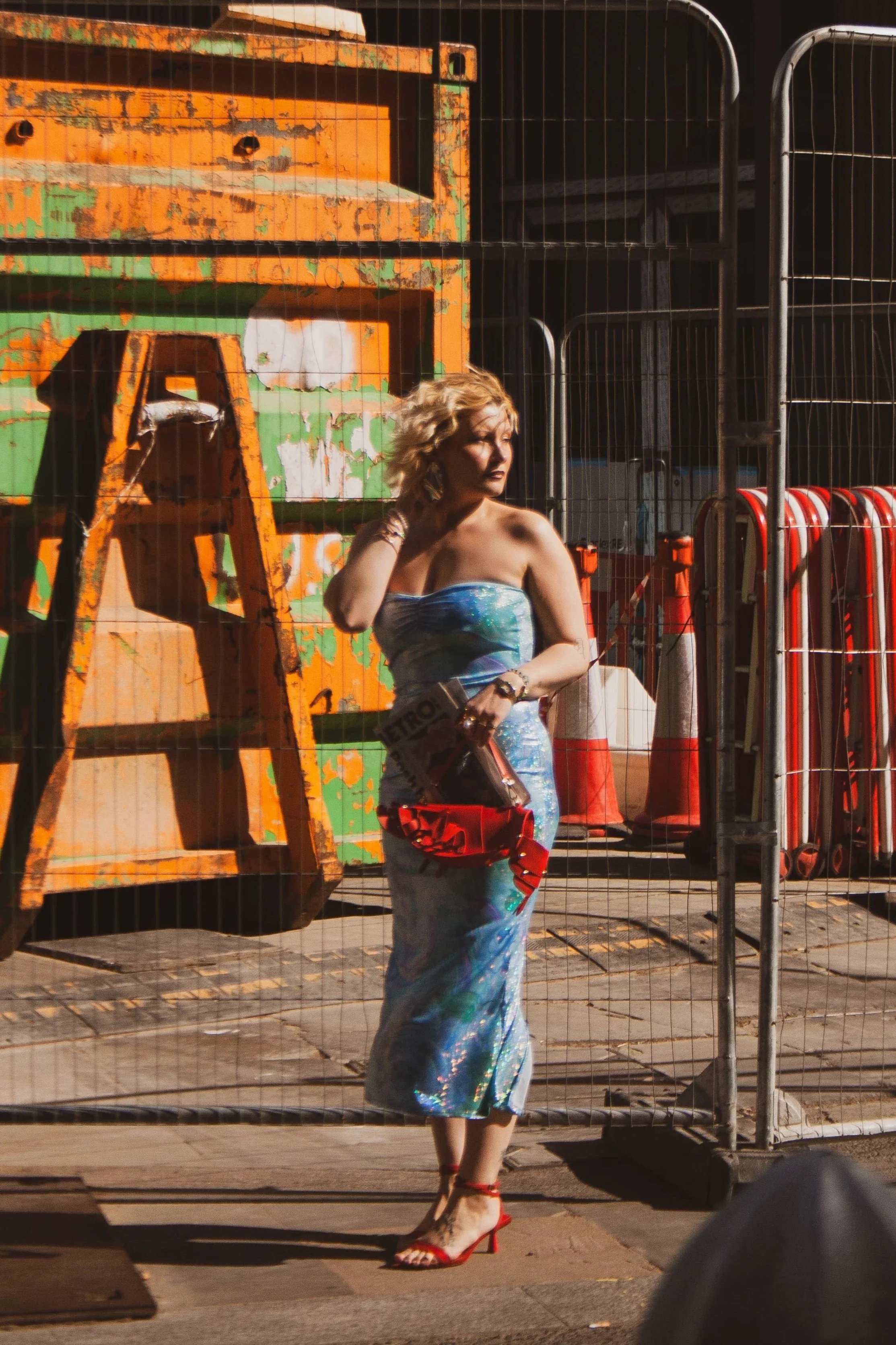 A woman with blonde curly hair wearing a colorful strapless dress and red high-heeled sandals standing behind a metal fence on an urban sidewalk. In the background, there are orange and green cargo containers, traffic cones, and metal railing.