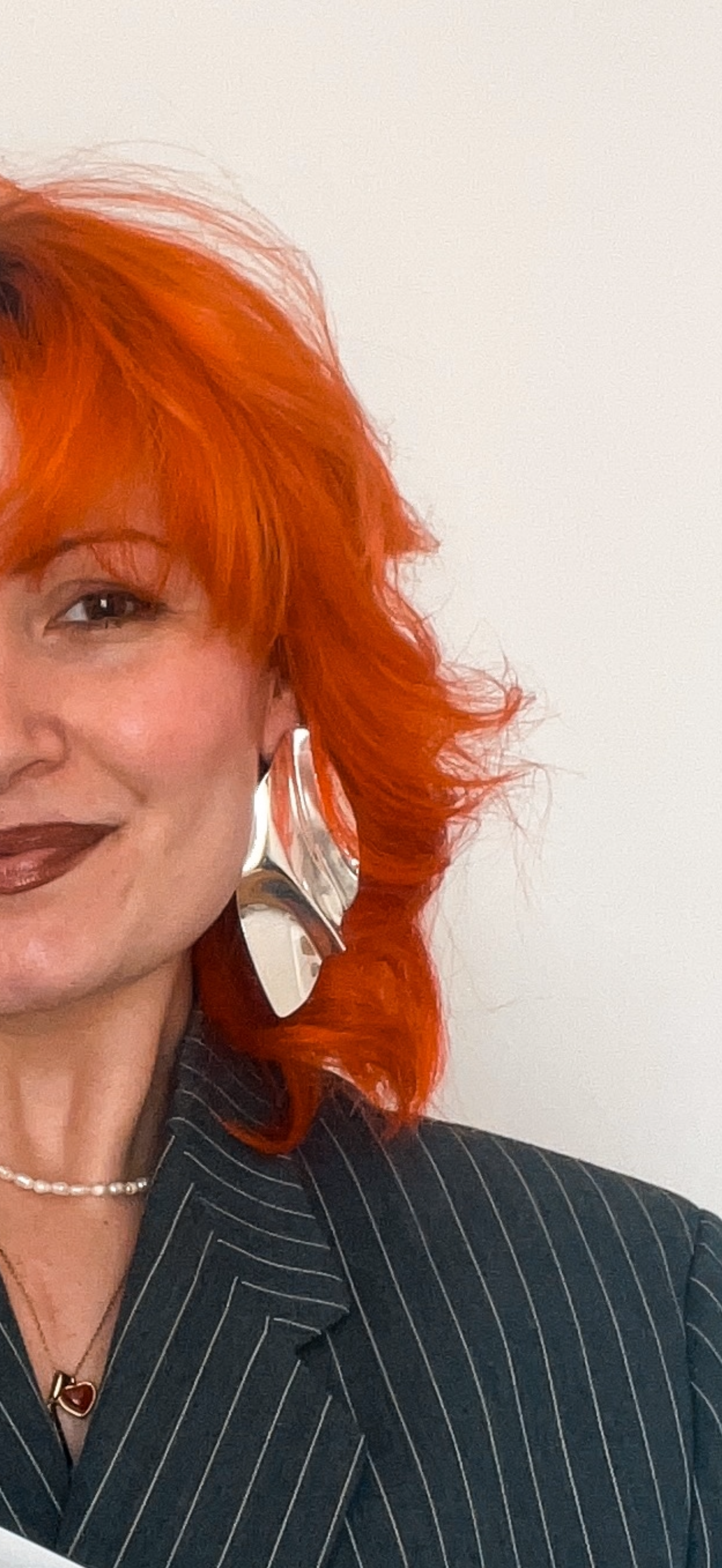 Close-up of a woman with bright orange curly hair, wearing a dark pinstripe blazer, a pearl necklace, and large silver earrings, smiling against a plain white background.
