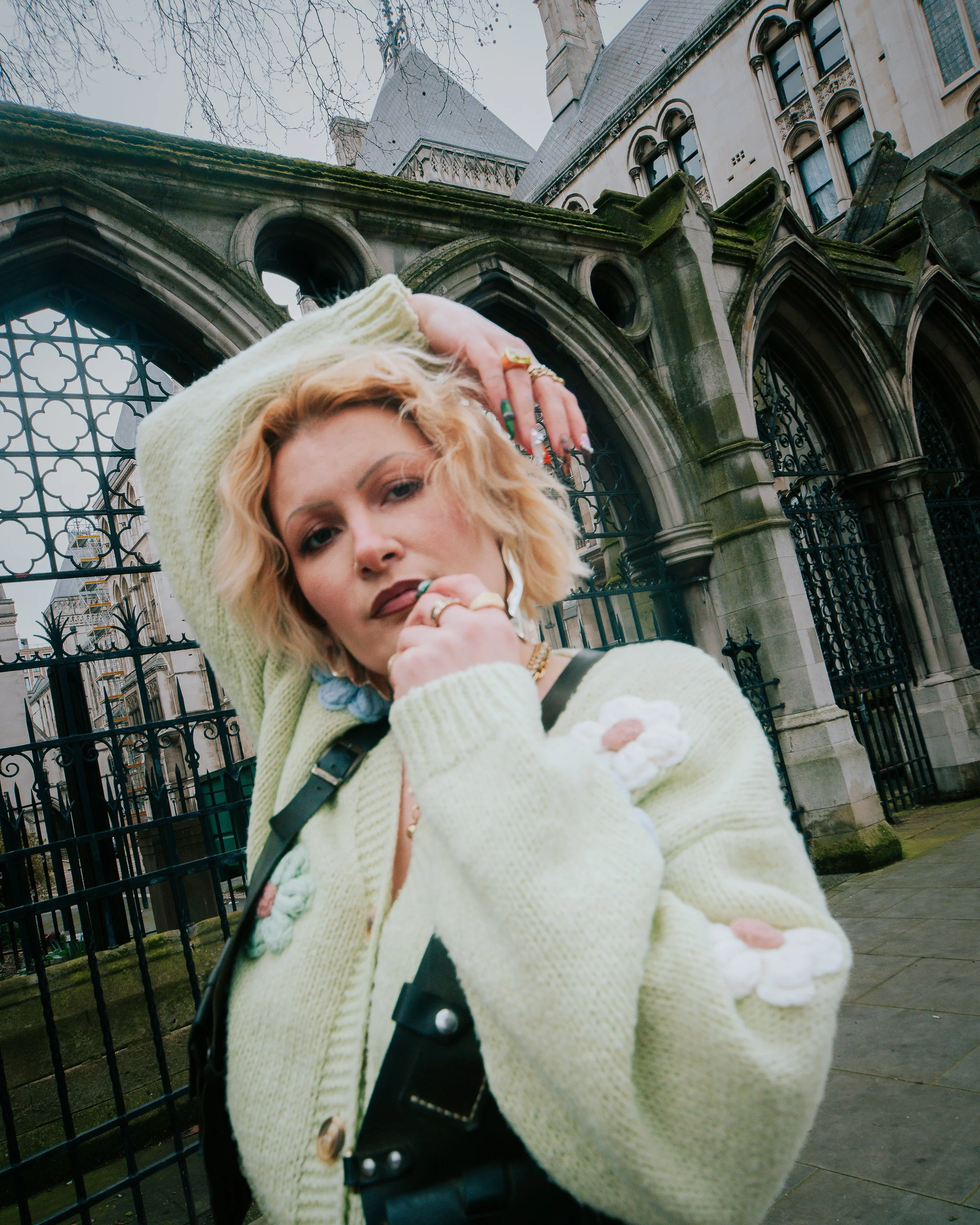 A woman with blonde curly hair and multiple rings wearing a cream-colored knitted cardigan and licking a flower-shaped ice cream cone, standing in front of a black wrought-iron gate in front of an old stone building with gothic architectural features and moss on the roof.