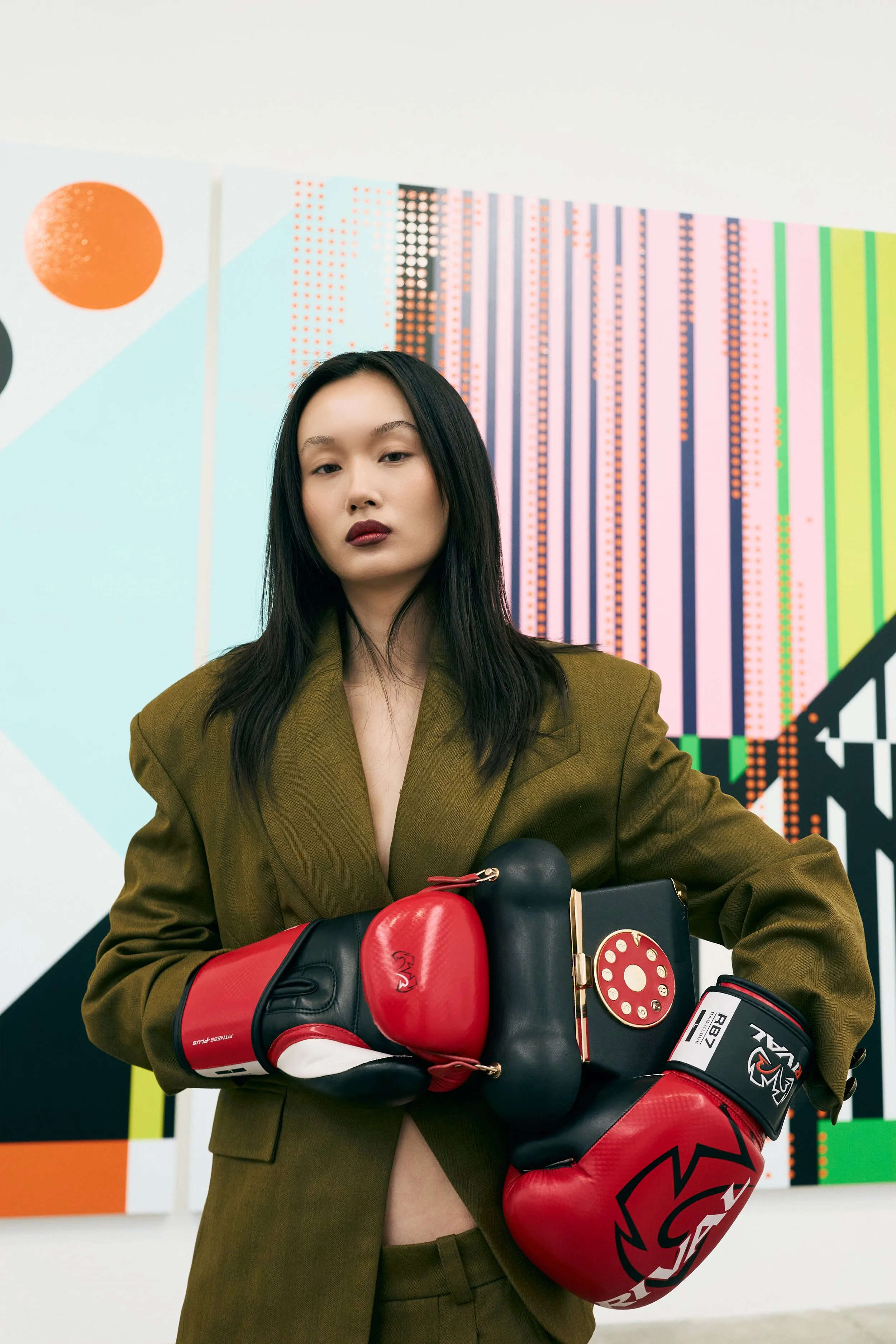 A young woman with long black hair wearing a brown blazer and boxing gloves, standing in front of a colorful abstract background.