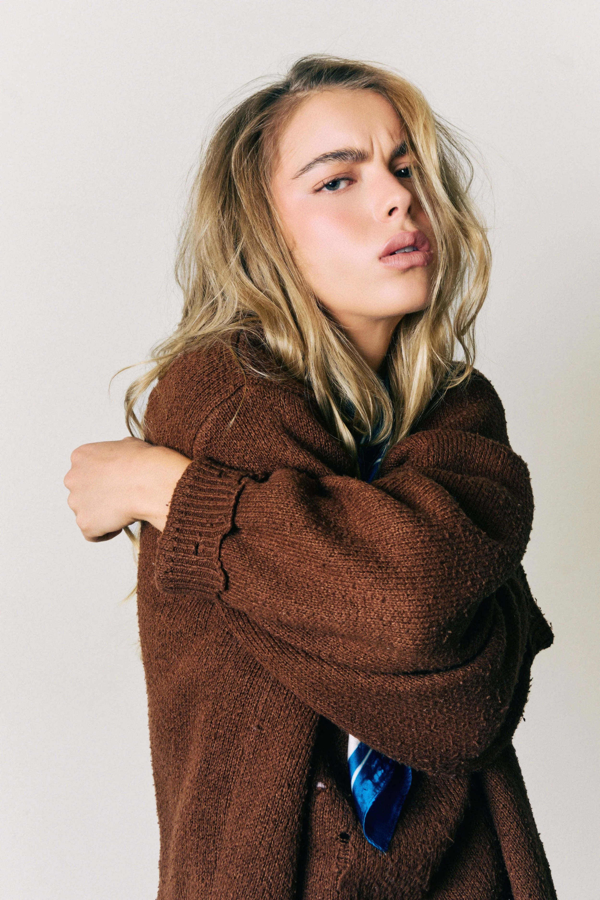 Close-up portrait of a young woman with wavy blonde hair, wearing a brown sweater, crossing her arms, with a serious facial expression against a plain background.
