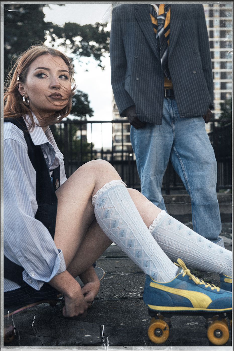 A young woman with red hair, wearing a striped shirt, knee-high socks, and roller skates, sitting on the ground with her legs extended. A person in a pinstripe blazer, blue jeans, and a tie stands behind her with hands in pockets. The scene appears to be outdoors in an urban area with buildings and trees in the background.