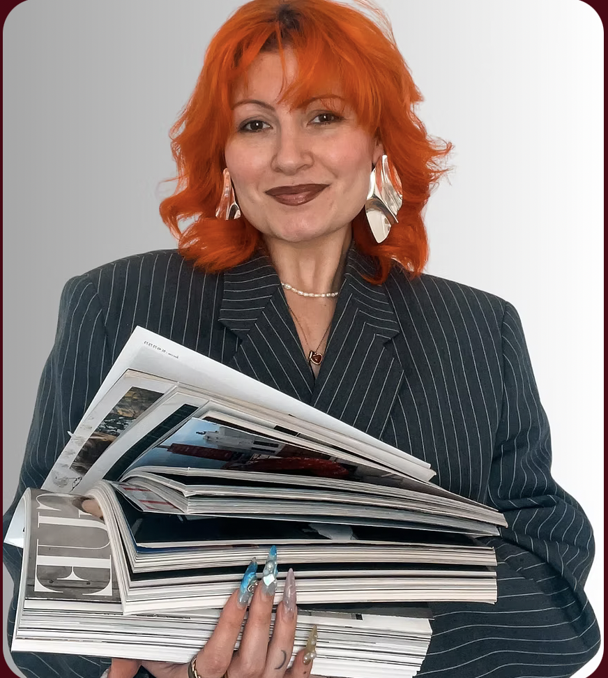 A woman with bright orange hair wearing a pinstripe blazer, large silver earrings, and jewelry, holding a stack of magazines and smiling.