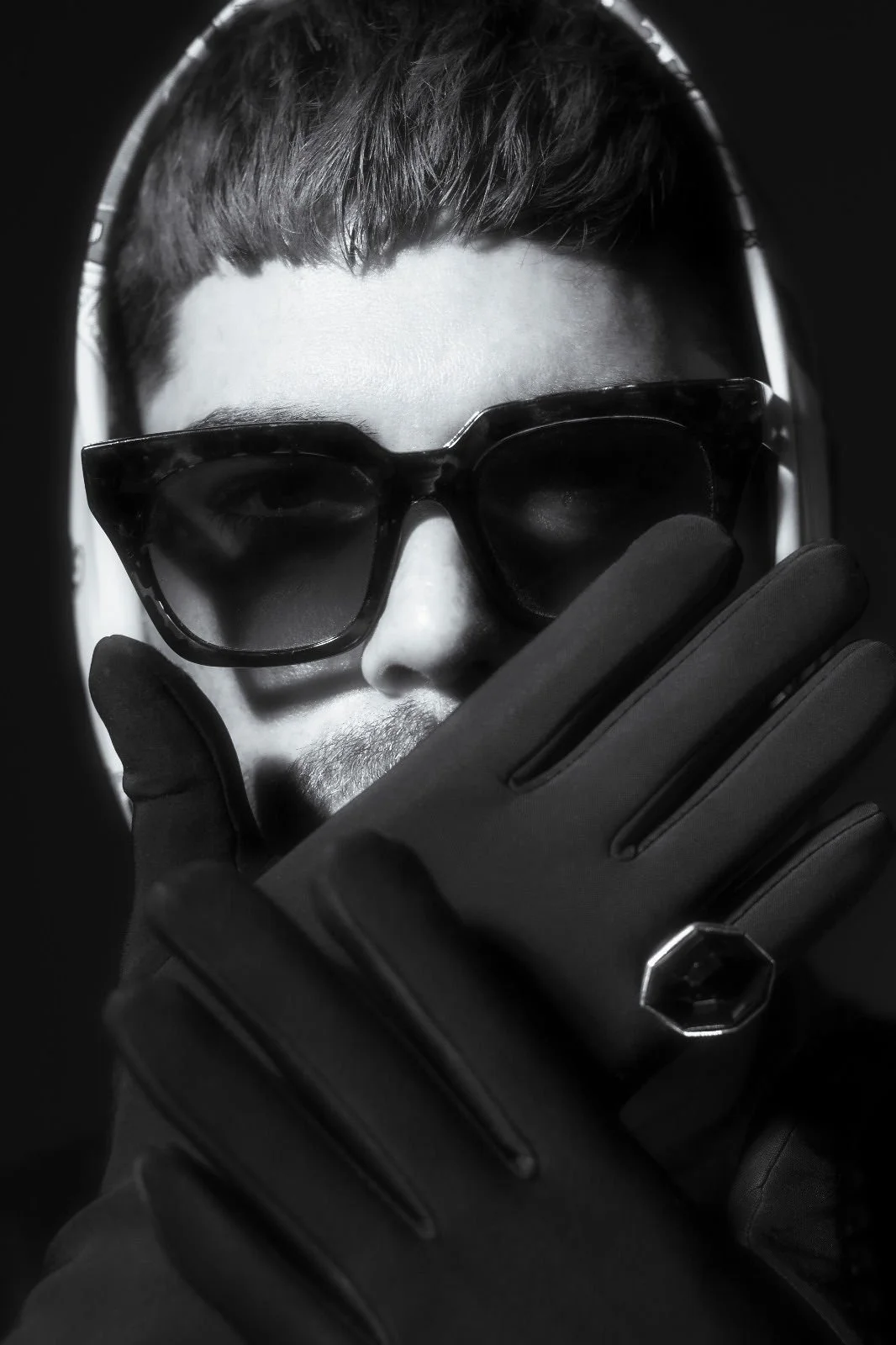 A monochrome close-up photo of a man with short hair wearing dark sunglasses, a headset, and gloves, touching his face with one hand.