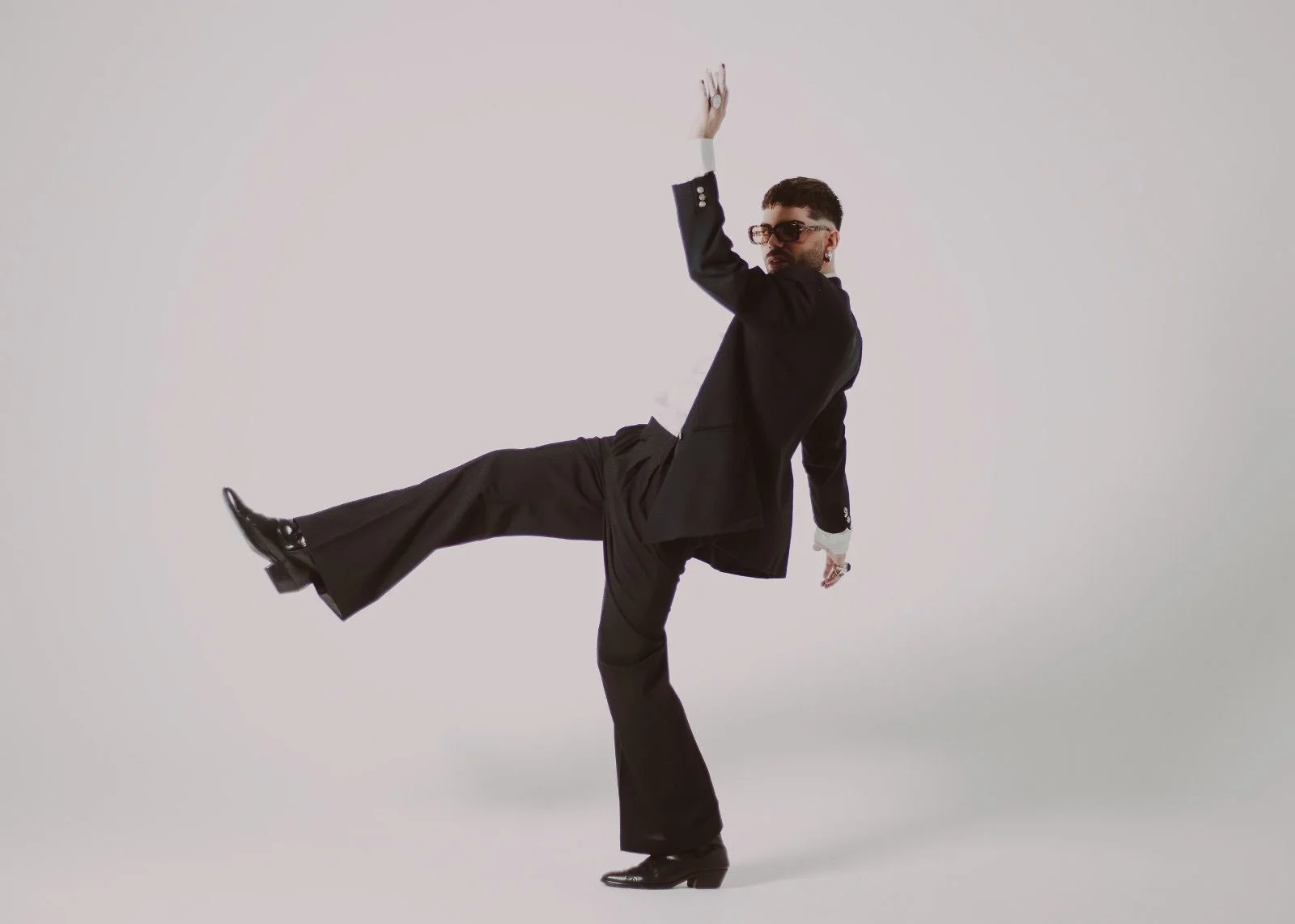 A man in a black suit and dress shoes strikes a pose with one leg extended sideways and one arm raised in the air against a plain white background.