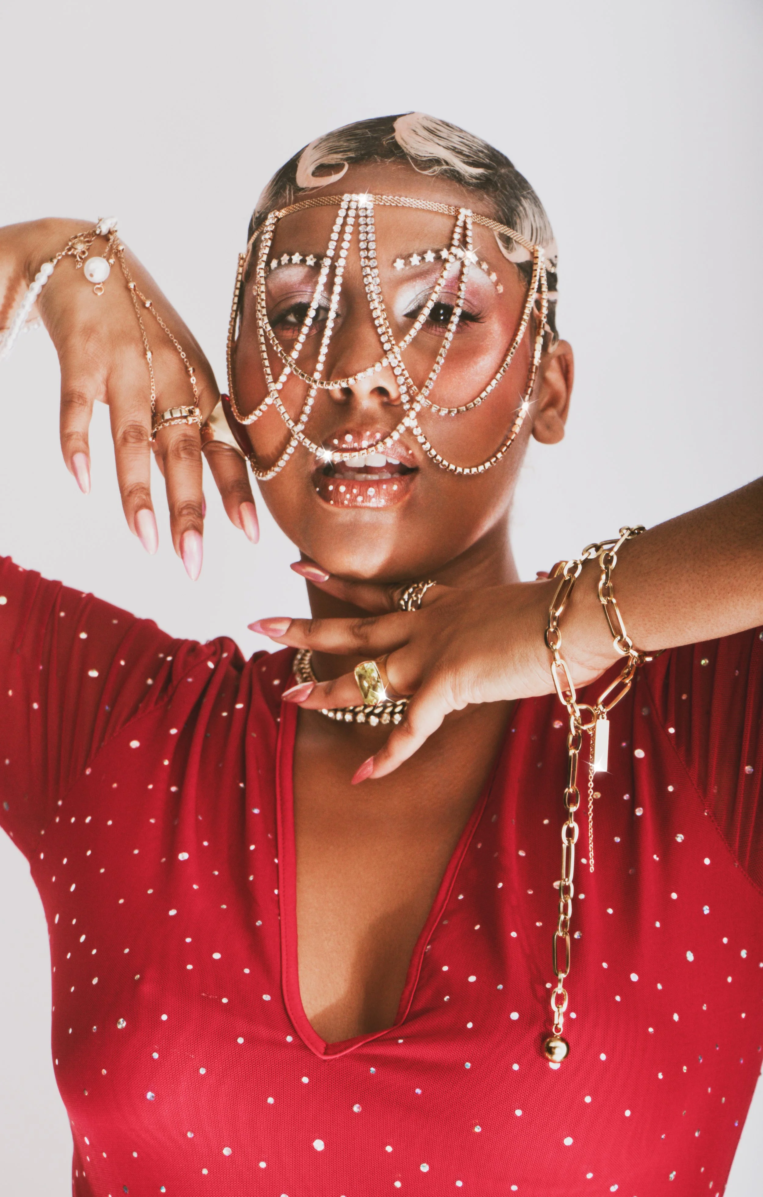 A woman with short hair wearing a red polka dot top, accessorized with gold jewelry, and an elaborate face chain covering her face, posing with her hands near her face against a plain background.