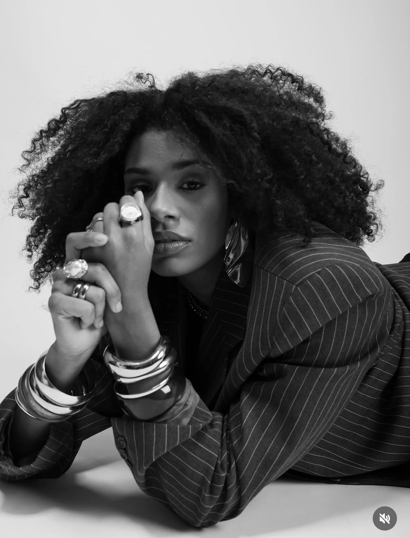 Black and white portrait of a woman with curly hair, wearing a pinstripe blazer, multiple bracelets and rings, resting her chin on her clasped hands.