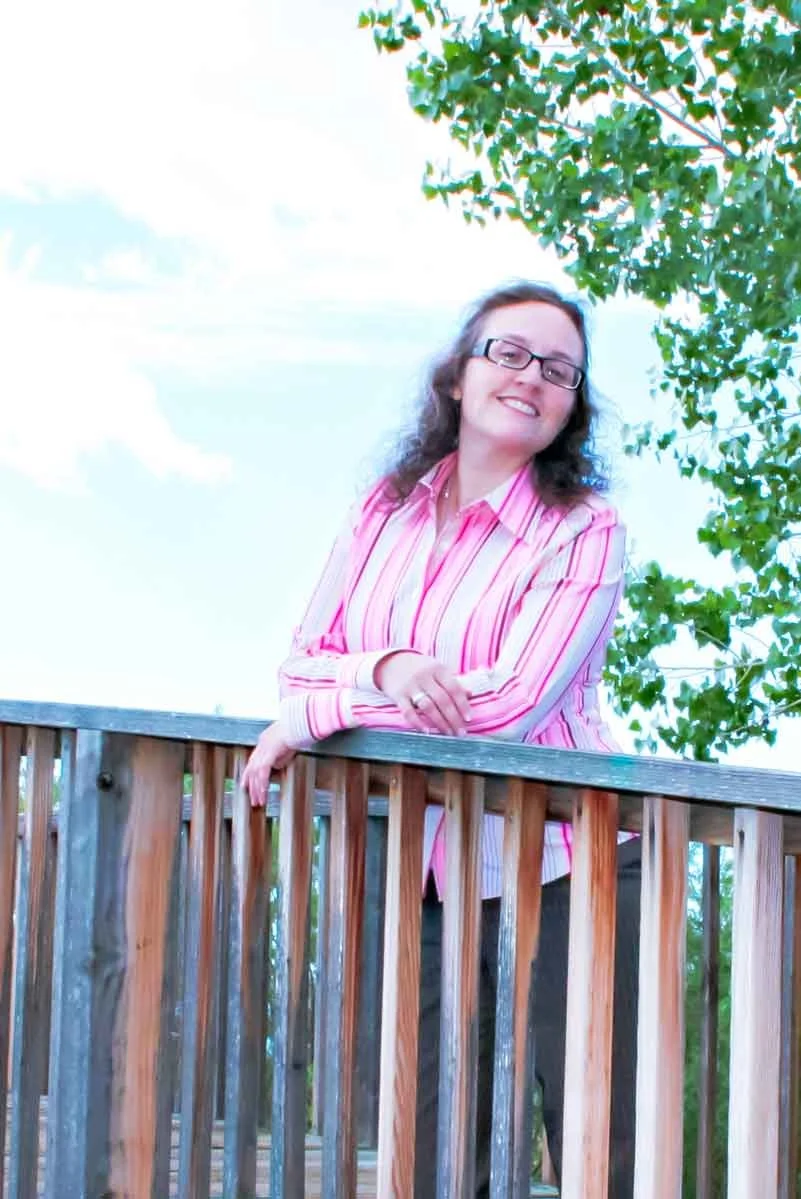 Lynne Marie O'Connor with glasses and curly hair, wearing a pink striped shirt, leaning on a wooden railing outdoors with green trees and a blue sky in the background.