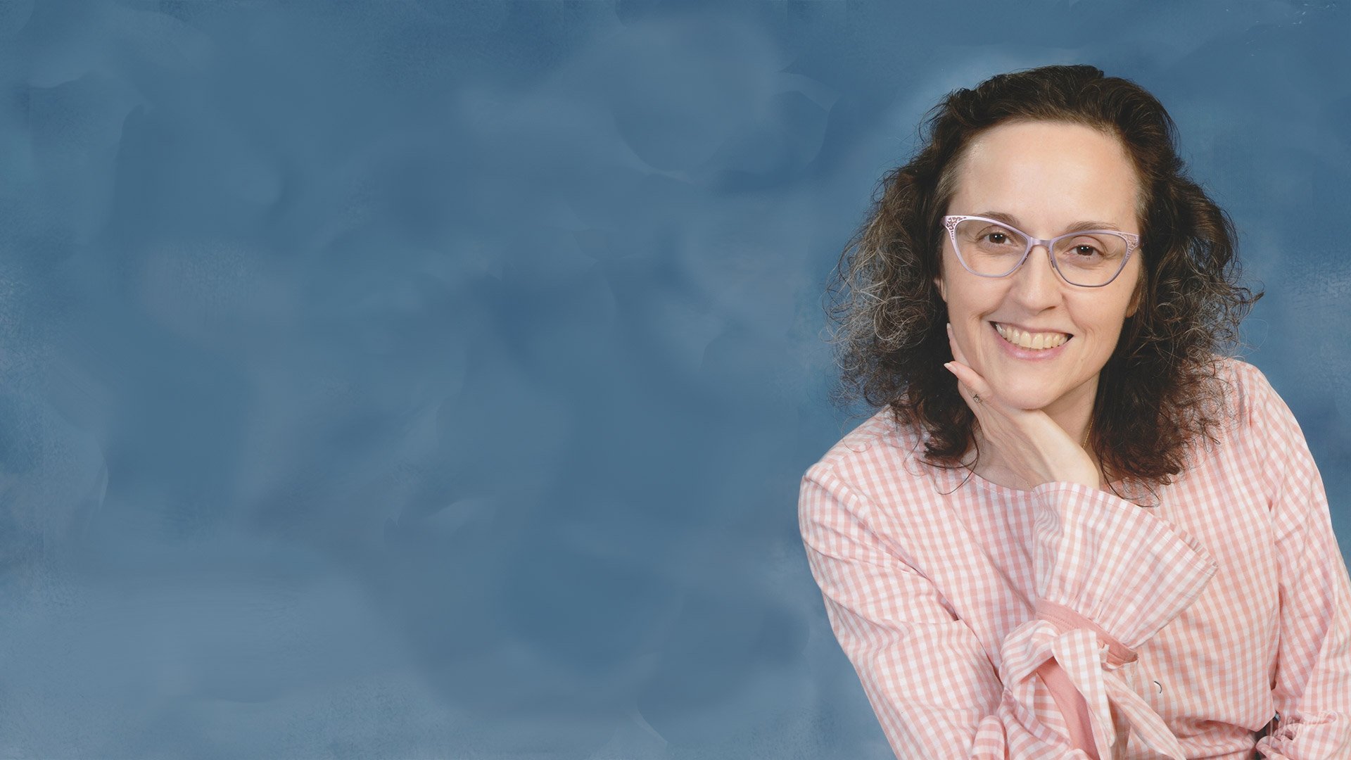 Lynne Marie O'Connor with curly dark hair and glasses smiling, resting her chin on her hand, wearing a pink and white checkered shirt, in front of a blue textured backdrop.