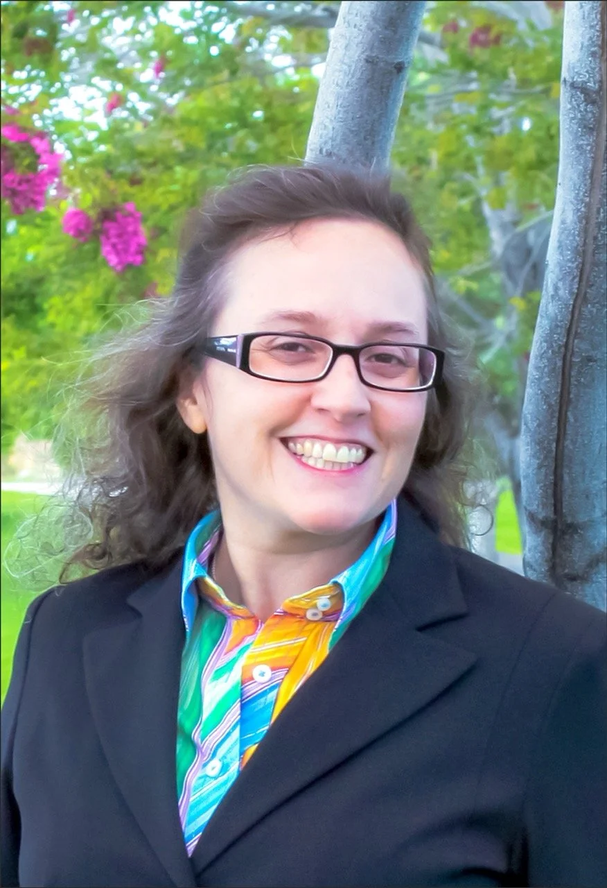 Lynne Marie O'Connor with glasses smiling outdoors, wearing a black blazer over a colorful, striped shirt, standing in front of a tree with pink flowers and green leaves.