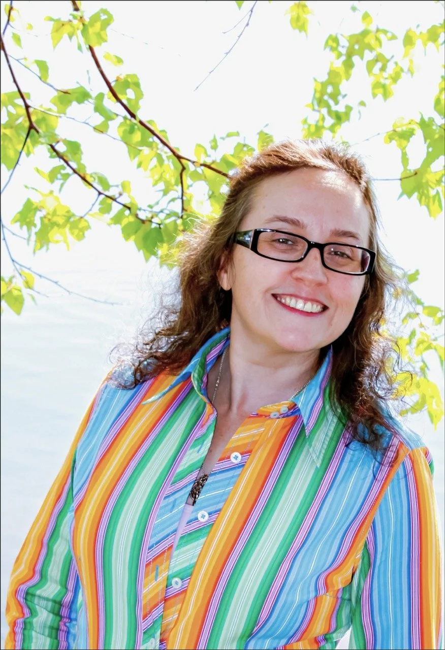 Smiling Lynne Marie O'Connor wearing glasses and a colorful striped shirt outdoors with green leaves and branches overhead.