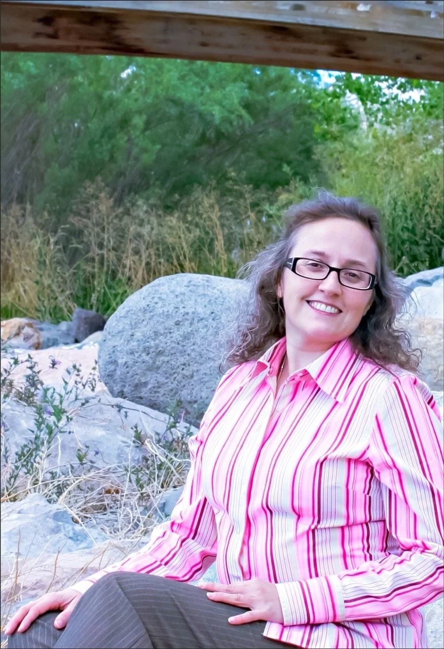 Lynne Marie O'Connor with curly hair and glasses smiling, sitting outdoors on rocks, wearing a pink striped shirt, with trees and rocks in the background.