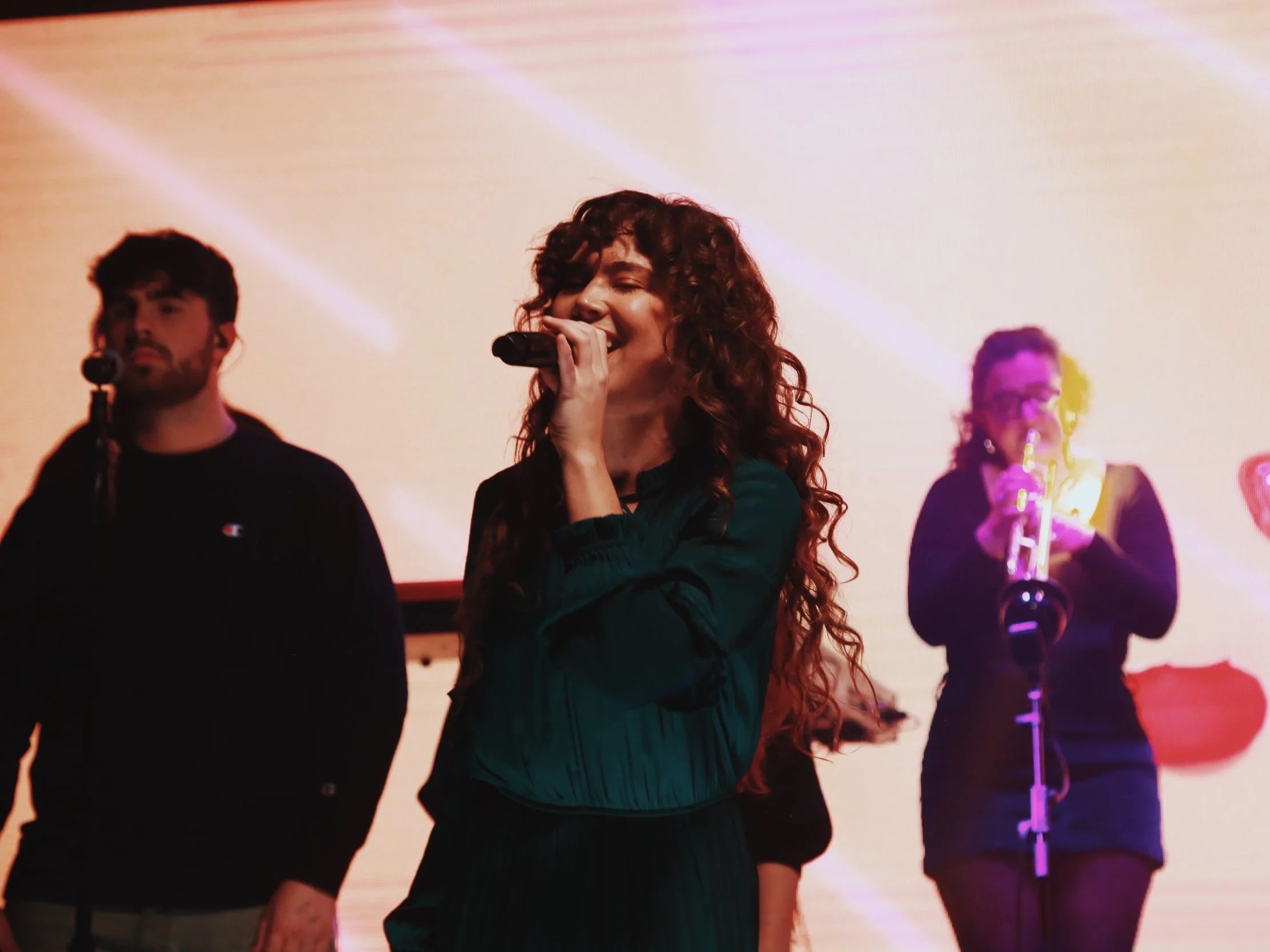 A woman with curly hair singing into a microphone on stage, with two other performers in the background, one playing a saxophone and the other holding a microphone.