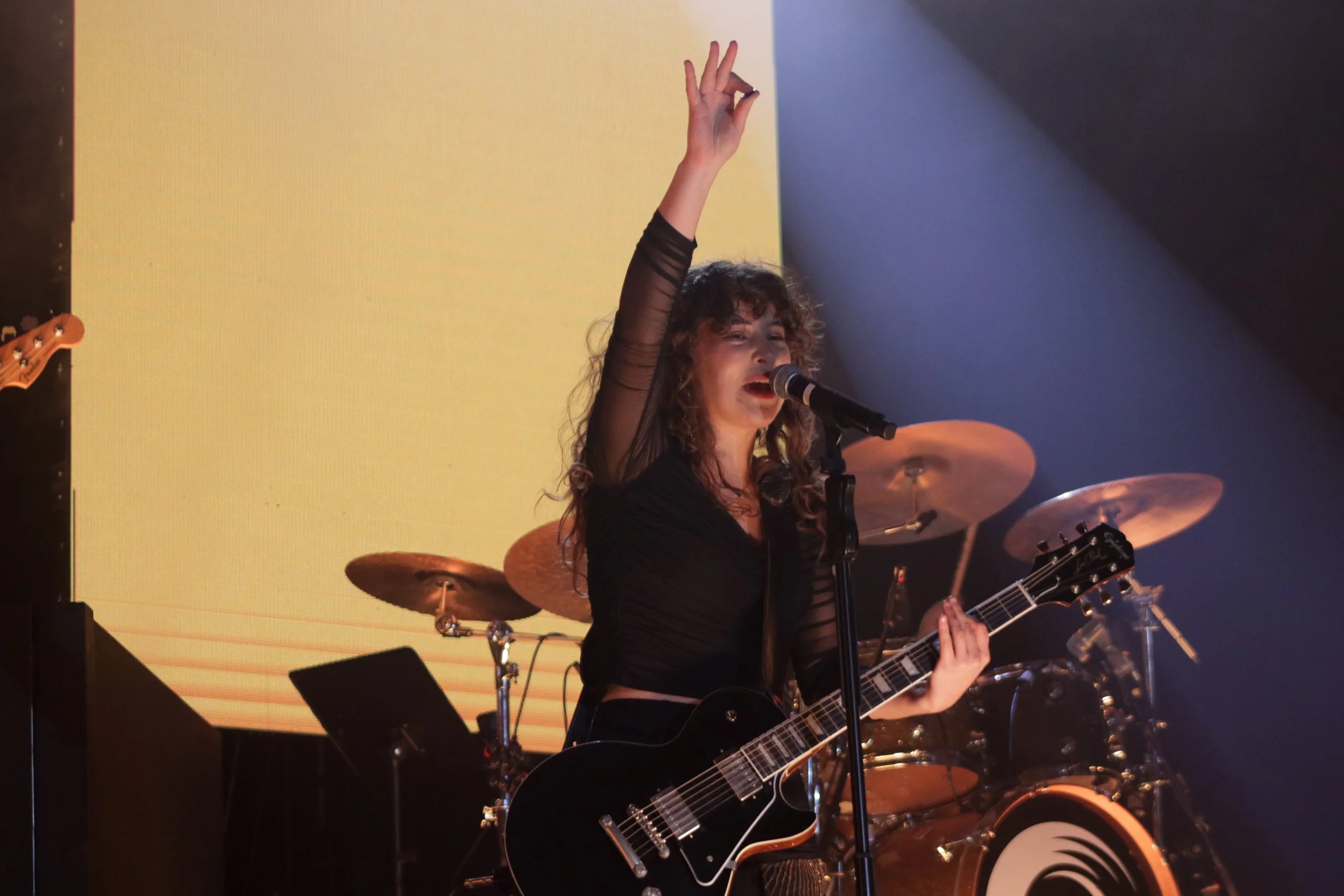 A woman with curly hair and a black sheer top is singing into a microphone and playing an electric guitar on stage. She has one hand raised in the air. There are drums behind her and a yellow-lit background.