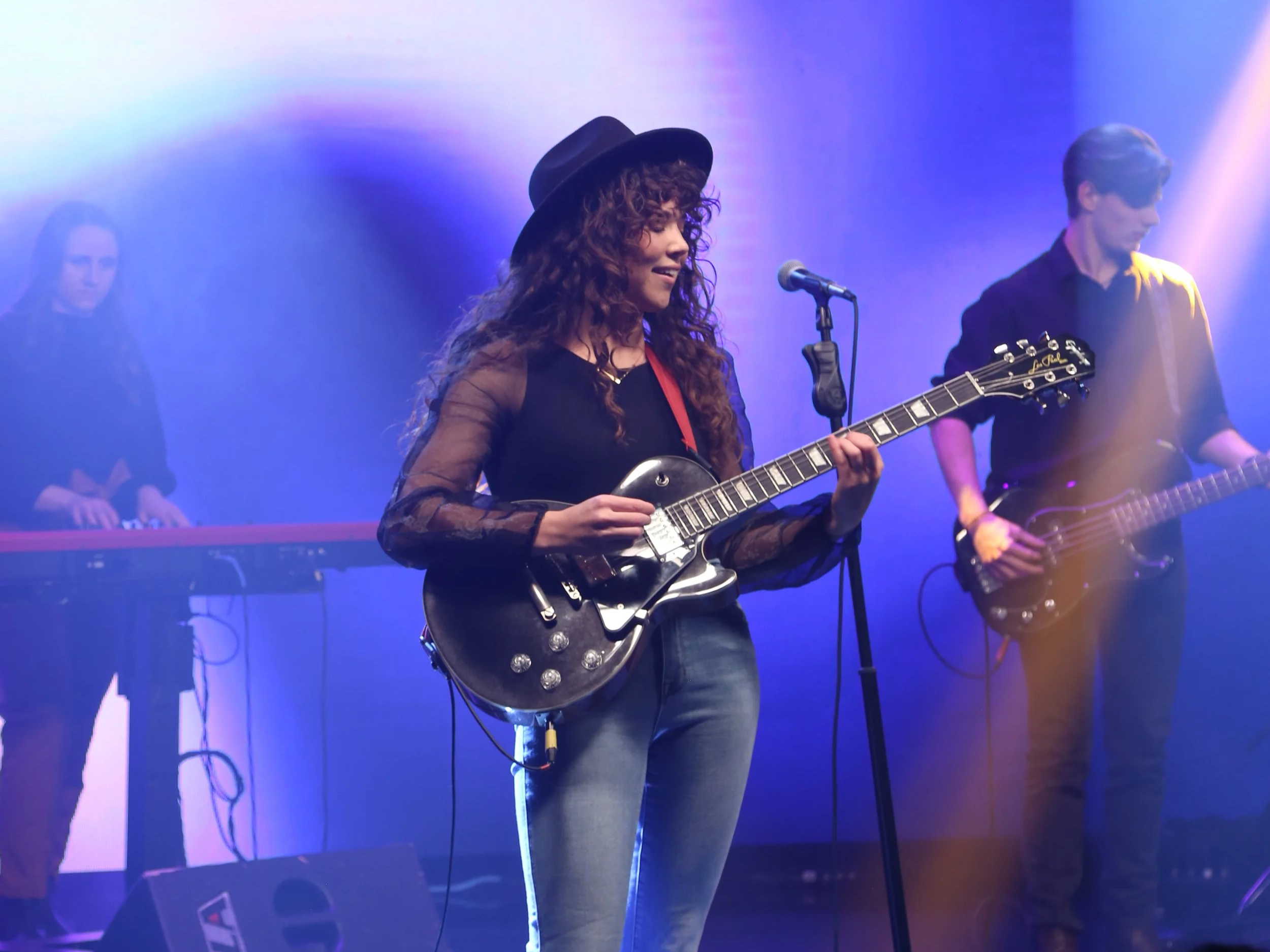 Musician with curly hair wearing a black hat and shirt, playing an electric guitar on stage with purple and blue lighting, and a keyboard player in the background.