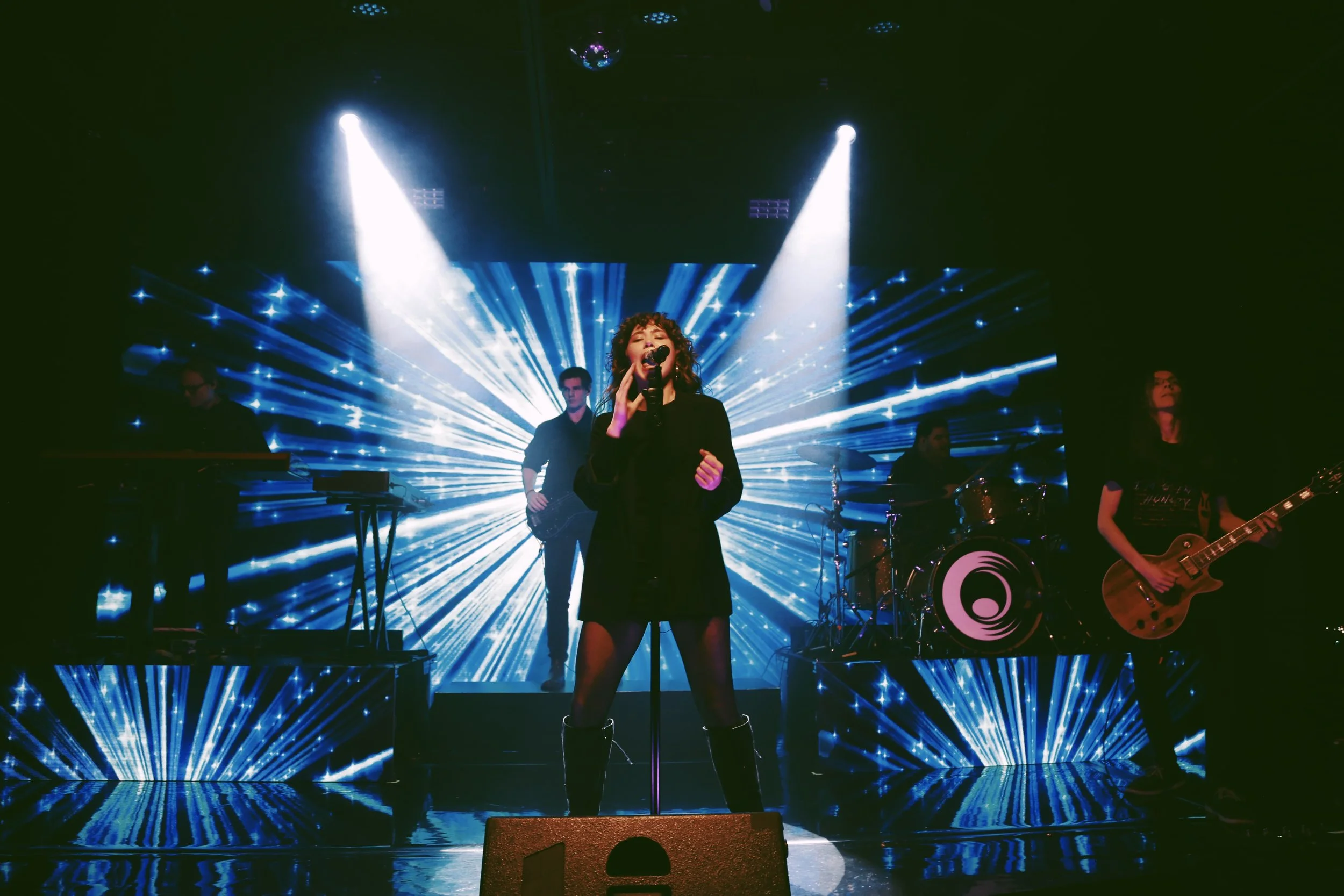 Female singer performing on a stage with vibrant laser lights radiating behind her, accompanied by musicians playing instruments.